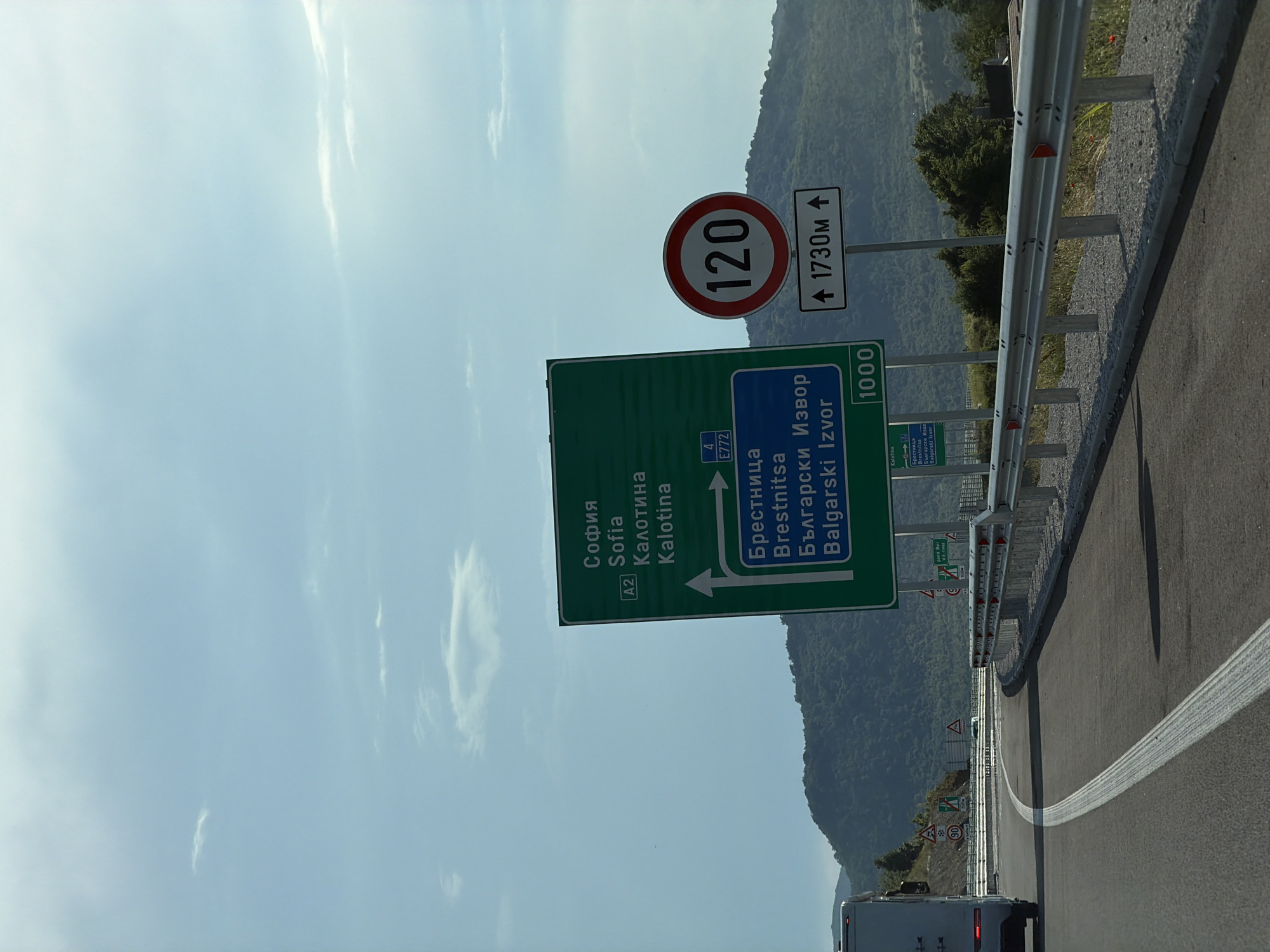 The image shows a road scene with a green directional sign indicating Sofia and Kalotina, as well as Brestnitsa and Bulgarski Izvor. A circular speed limit sign displays "120" and a supplementary sign indicates "1730m". The road curves to the right, with a metal guardrail along the side. In the background, a lush, tree-covered mountainside is visible under a partly cloudy sky. The location is identified as Peshtera, Bulgaria, however, there is no visual information in the image to confirm this specific location. The scene appears to be on a highway or major road.