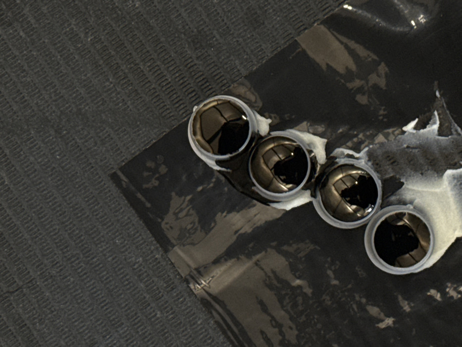 Four dark, highly reflective, spherical objects, approximately 1.5 cm in diameter, are diagonally aligned on a shiny black plastic sheet. Each sphere is encircled by an irregular, light grey, semi-solid substance, possibly an adhesive or sealant. The black plastic sheet rests upon a darker grey, coarsely textured surface, likely a mat or fabric. The spherical objects reflect an interior architectural space featuring bright overhead lighting and distinct arched structures, possibly ceilings or large windows. Reflections also show the silhouette of a person holding a device (likely the photographer's hand and phone/camera) and a partial reflection of another individual. The image does not contain sufficient visual cues to forensically identify a specific location within Moscow, Russia. The reflections indicate a modern indoor environment.