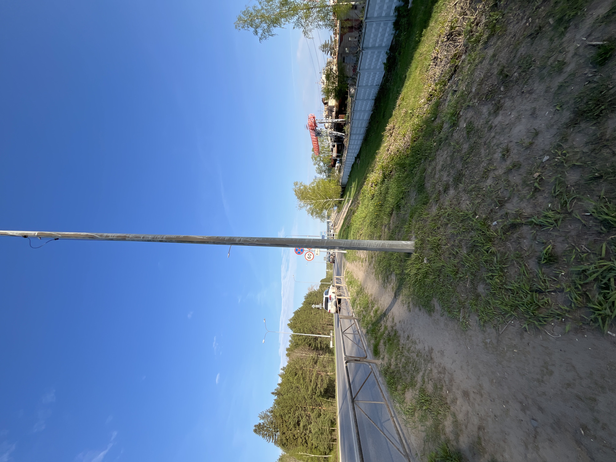 The image depicts a street scene in Chaikovsky, Russia. A metal guardrail separates the road from a tree-lined area on the left. On the right, a dirt path runs parallel to the road, with a grassy area and a fence behind it. A speed limit sign is visible in the distance, indicating a 60 km/h limit. Several cars are on the road. The sky is clear and blue.