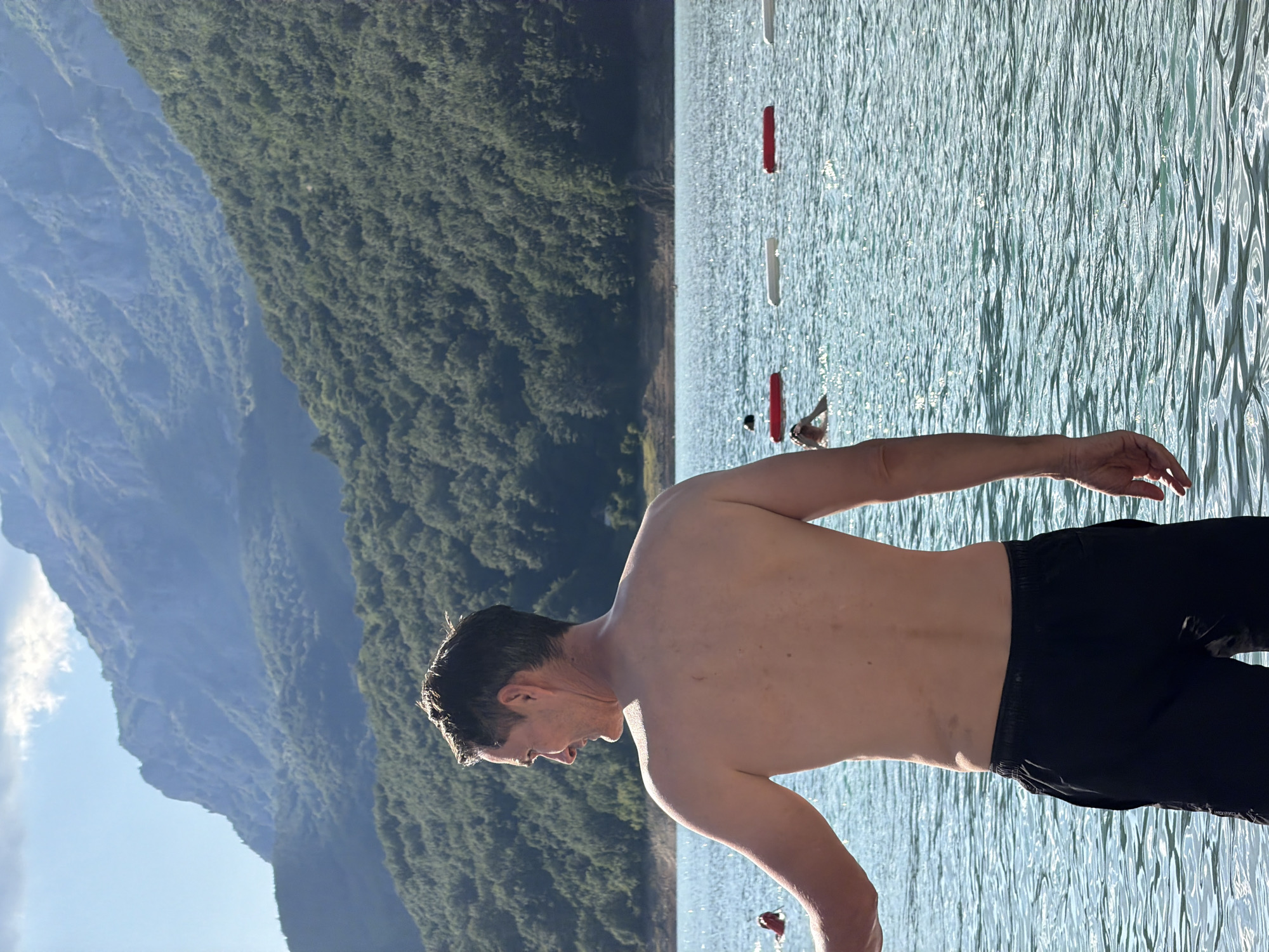 A man, shirtless and wearing black shorts, stands in the clear, blue water of a lake, facing away from the camera and looking to the left. His skin is tanned, and he has short, dark hair. The lake is populated by a few other swimmers in the distance. Across the water, a lush, green, tree-covered mountain range rises, its peaks partially obscured by a hazy sky. The scene is lit by bright sunlight, casting reflections on the water. The location is identified as the Piva Lake in Plužine, Montenegro.