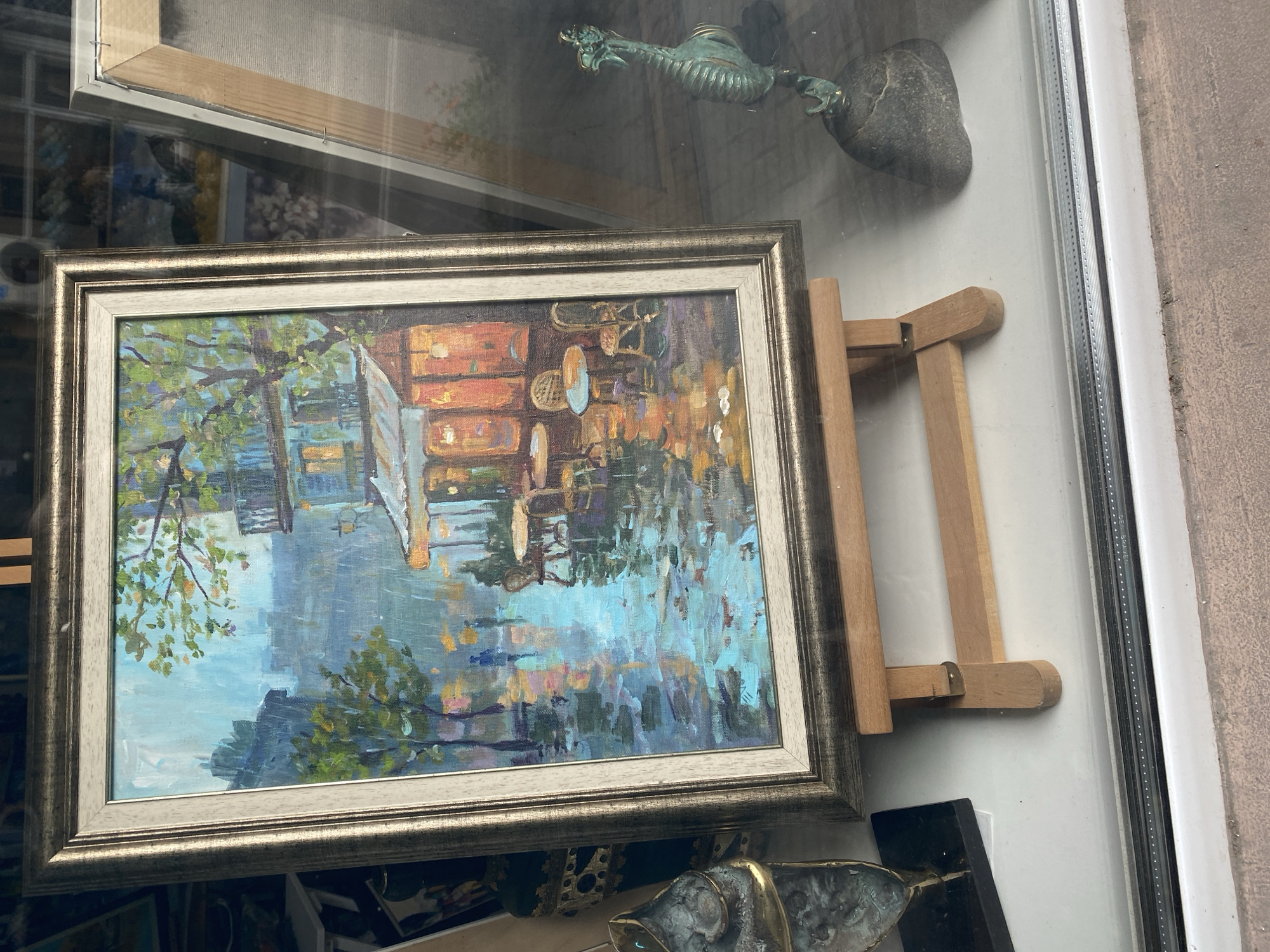 The image is a close-up shot of a framed painting displayed in a shop window in Varna, Bulgaria. The painting depicts a landscape scene with water, trees, and a building. The frame is silver-colored and ornate. The painting rests on a small wooden easel. Above the painting, visible through the glass of the shop window, is a metal figurine of a dragon or sea serpent, and a smooth, grey stone. Below the painting are other objects partially visible. The window frame is white. The overall scene has a slightly cluttered, artistic feel.