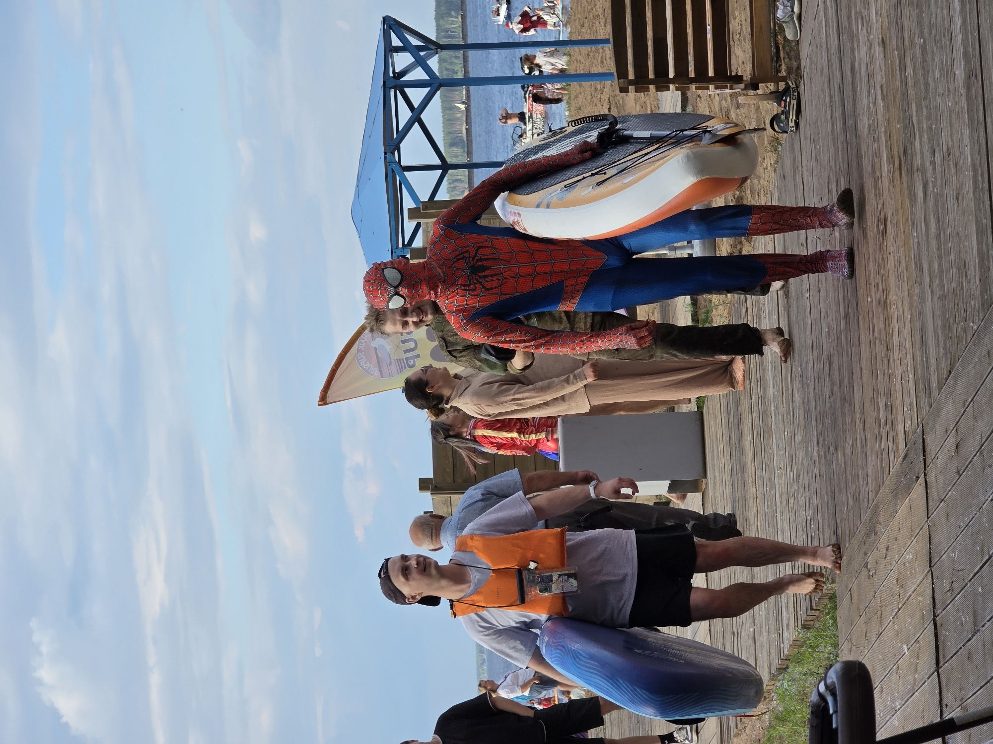 On a wooden boardwalk near a body of water, a person dressed as Spider-Man walks carrying a paddleboard. The boardwalk is lined with grass and leads towards a sandy beach. In the background, people are seen near the water, some with watercraft. A blue canopy structure is visible on the beach. The sky is partly cloudy. The scene appears to be at a recreational area in Chaikovsky, Russia.