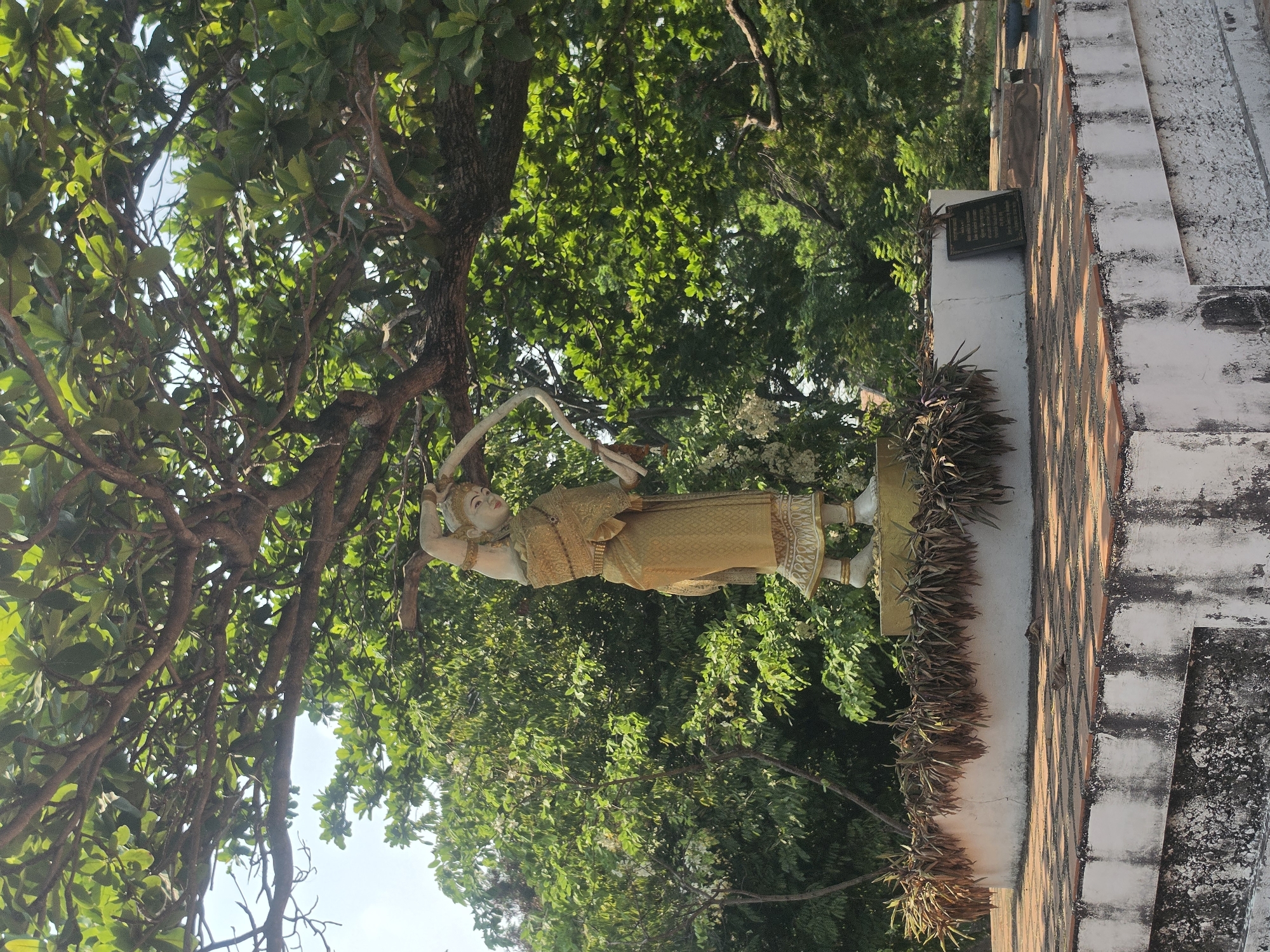 The image depicts a statue of a person in traditional Thai clothing, positioned on a structure that appears to be part of a temple or historical site. The statue is set against a backdrop of lush green trees. The location is Phra Nakhon Si Ayutthaya City Municipality, Thailand. The structure supporting the statue is made of brick and white stone. The sky is visible through the trees.