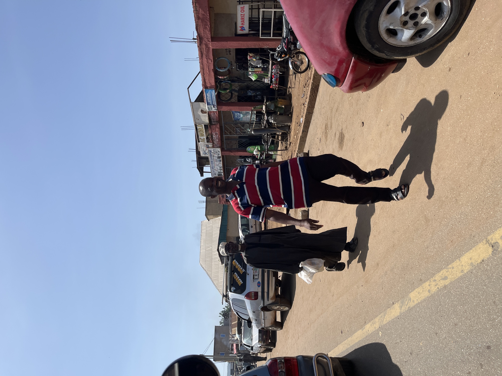 This image captures a vibrant street scene in Rafin Par, Nigeria, under a clear blue sky. The picture is taken on a bright, sunny day, indicated by the strong sunlight and prominent, elongated shadows cast on the light-colored paved road, suggesting either morning or late afternoon.
In the foreground, a man wearing a red, white, and blue striped polo shirt and dark trousers, sporting sandals, stands smiling directly towards the camera. His long shadow stretches behind him on the dusty ground. Adjacent to him, a person dressed in a dark robe and a white cap is visible, partially obscured, standing near a parked silver SUV-like vehicle. The vehicle has text on its rear window, including "BIG MAN". The front fender and wheel of a red car are visible in the extreme foreground, suggesting the image might have been taken from within a vehicle.
On the right side of the frame, a two-story commercial building lines the street. Its facade is reddish-brown, and the ground floor features several open-front shops displaying various goods, possibly auto parts or hardware, including tires and tools. Visible signs on the building include "HARIZ OIL" at the top, and partially legible signs for "CHARLES" and "AC WORLD". A motorcycle is parked near the building. Further down the street, additional vehicles are parked, and traces of a faded yellow road line are visible on the asphalt. The setting conveys a bustling, everyday urban or semi-urban environment.