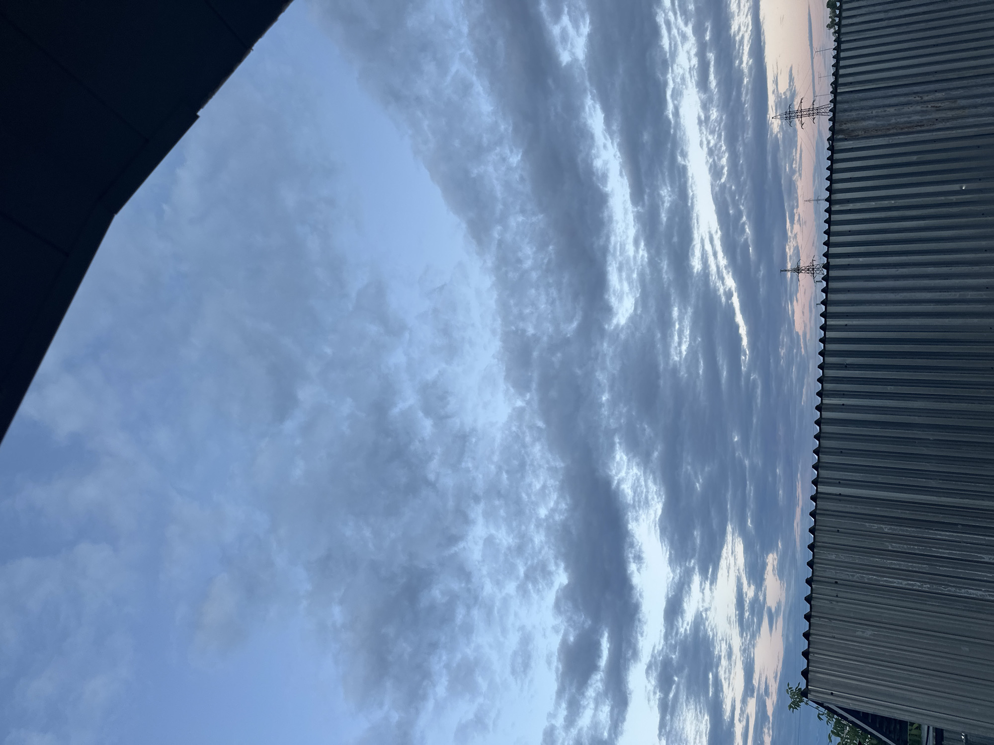 The image depicts a sky with rows of stratus clouds. In the top right corner, a galvanized steel corrugated roof is visible, and in the background, power lines are visible. A portion of a dark roofline appears in the upper left corner of the image. The image has a slight sideways tilt. It is daytime, likely at dawn or dusk. The location is possibly Yerevan, Armenia, based on the presence of power lines and a corrugated roof. There are no people visible in the image.