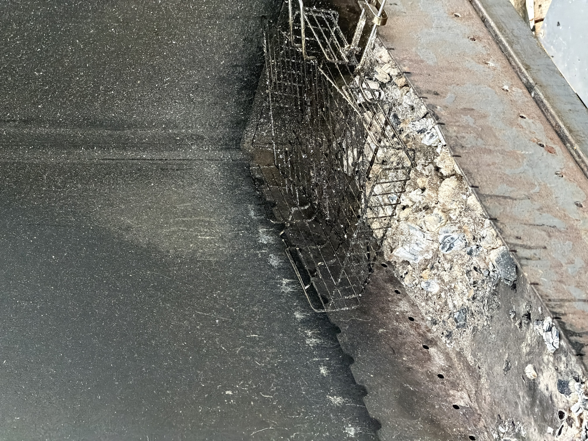 The image shows the interior of a used barbecue grill, likely located in a yard or outdoor recreational area in Bol'shaya Ven'ya, Russia. The grill is filled with ash and debris, remnants of past fires. A metal grill rack, heavily coated with grease and soot, is positioned above the ash. The interior walls of the grill are also blackened and speckled with ash particles. The overall scene suggests a state of disuse and the need for cleaning. There are no people or animals visible in the image.