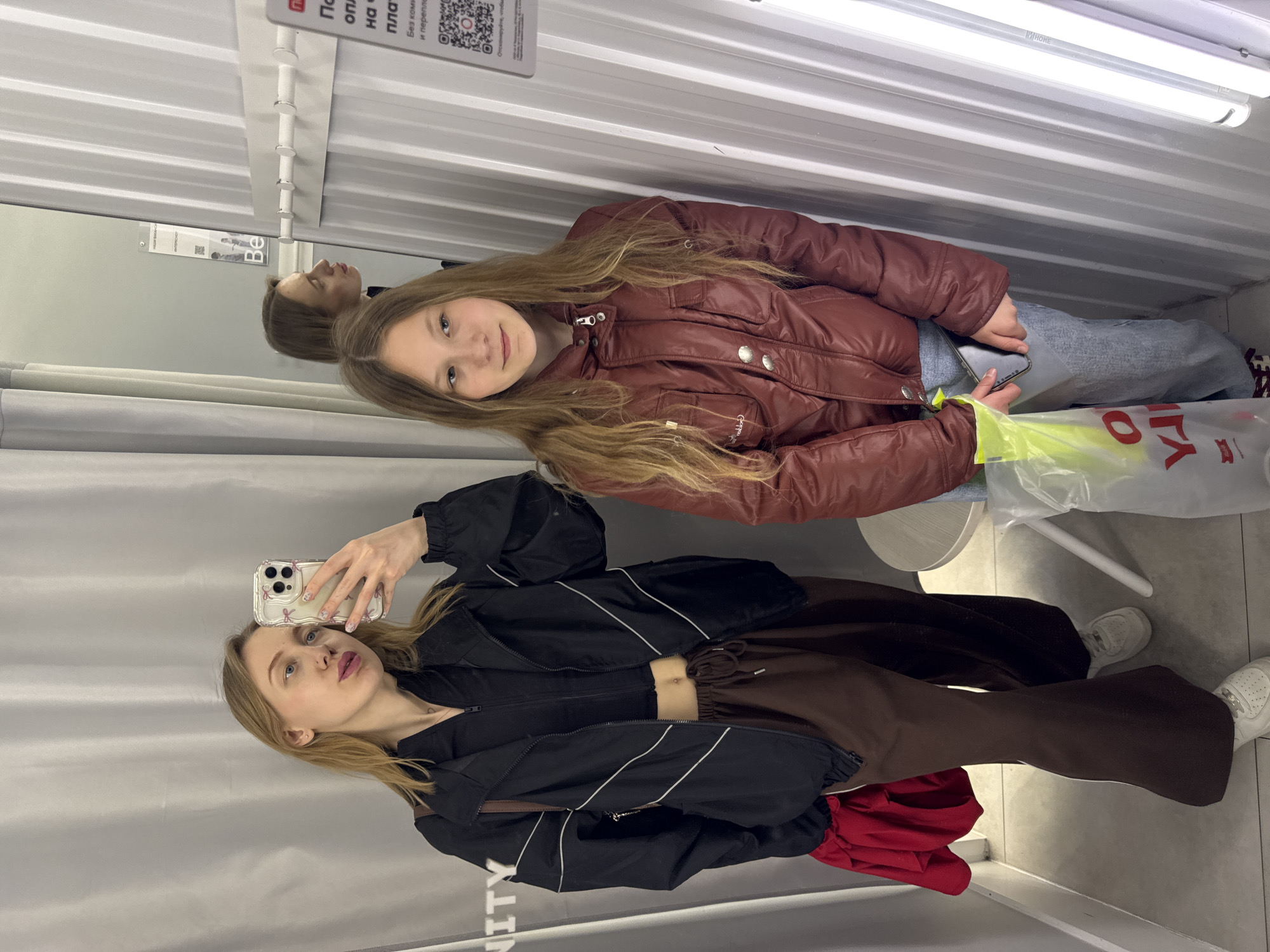 Here is a forensic description of the image.
**Scene:**
The image appears to be taken inside a fitting room, possibly in a retail clothing store. The walls of the fitting room are white, with a horizontal grooved pattern. There's a mirror in the fitting room reflecting one of the individuals. Overhead, there is a fluorescent light fixture providing illumination. A sign with text in Russian is visible near the top of the frame.
**People:**
Two female individuals are present. One is possibly taking a selfie with a smartphone, as it is being held up in front of her face. The other individual is beside her.
**Subjects:**
Clothing is a primary subject of the image, as it is being tried on or displayed.
**Objects:**
Several objects are discernible:
- A smartphone with a decorative case.
- Articles of clothing on the individuals, including a dark jacket, brown pants and a red jacket.
- A shopping bag.
- A small white stool
**Actions:**
The individual with the phone is taking a picture, while the other is posing or simply present in the shot.
**Interactions:**
The two individuals appear to be together, possibly shopping.
**Environment:**
The environment is a typical fitting room with basic furnishings and lighting. The location is likely a store.
It is NOT possible to confirm the location is in the city of Ovoshchnoy, Russia, from the image.