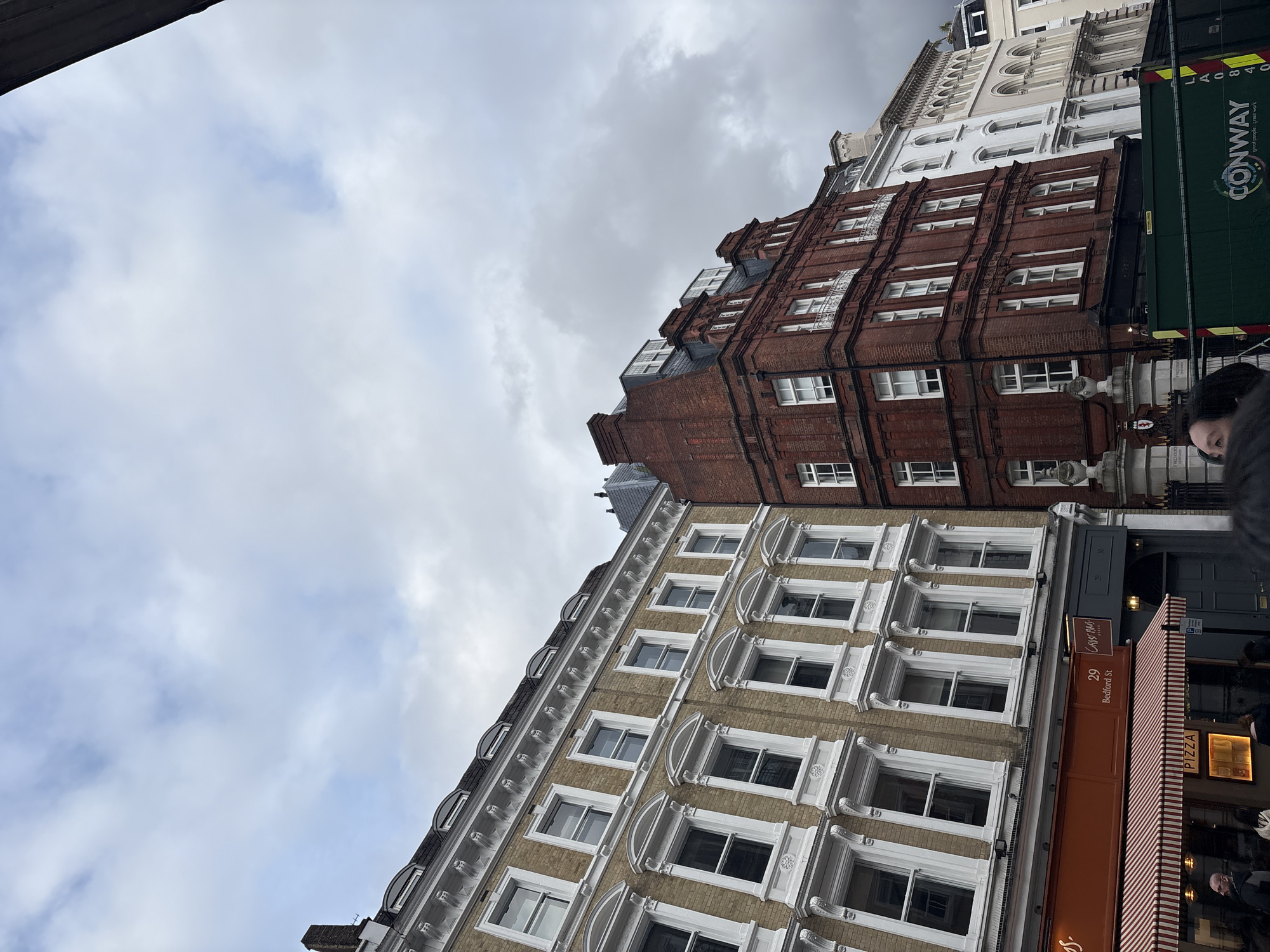The image shows a street scene in London, United Kingdom. Looking upwards at an angle, several buildings are visible. The nearest building has a beige facade with white window frames. A business called "Crust Bros Pizza" is located at 29 Bedford Street on the ground floor. Next to it is a red brick building with white-framed windows. Adjacent to this building is a bus or large vehicle with "CONWAY" printed on it. The sky above is cloudy. A few people are visible in the lower right corner.