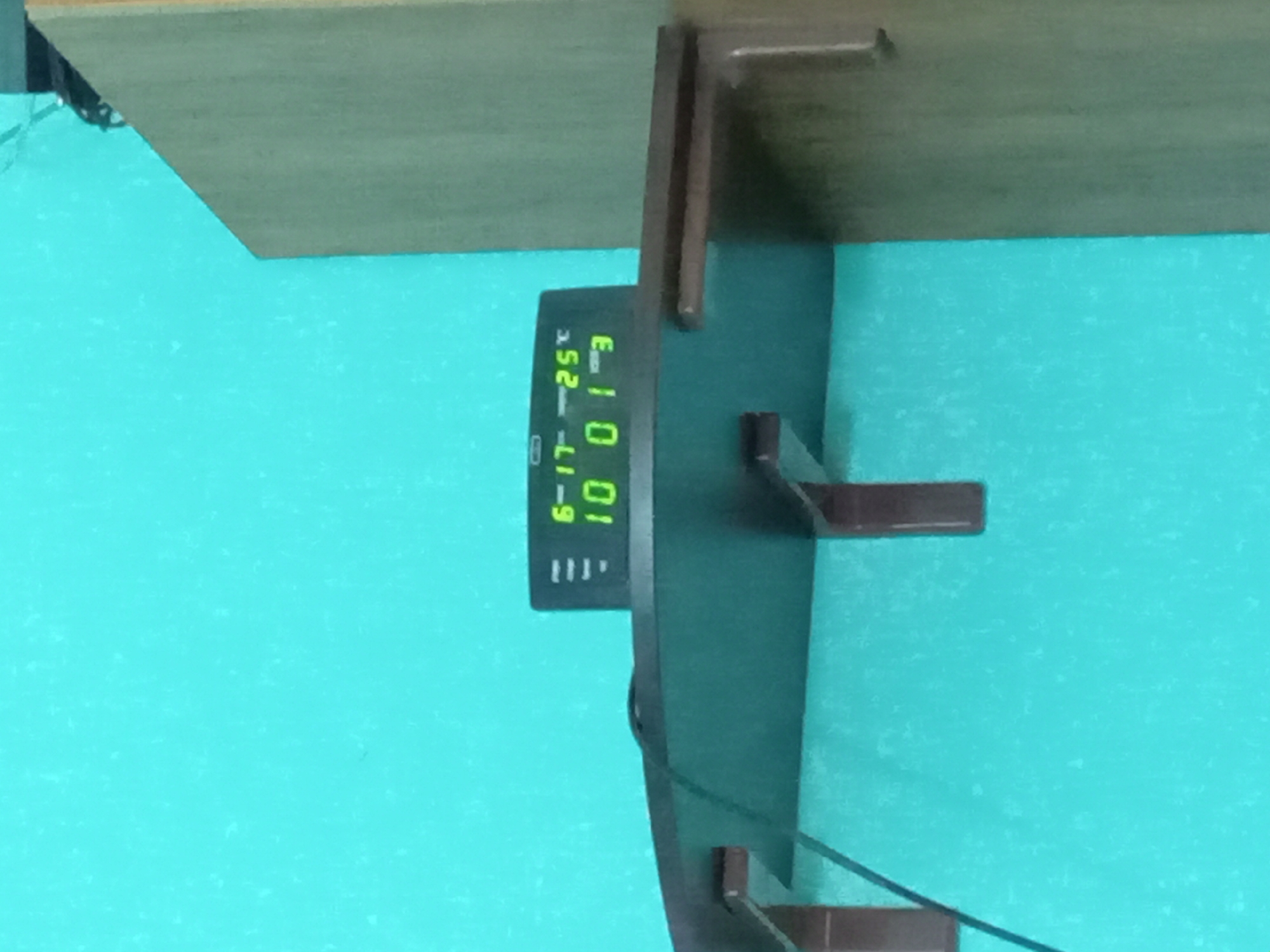 The image shows a close-up of a digital display, likely from a clock or thermometer, mounted on a dark green surface. The display shows green digital numbers and text. Visible text includes "E", "-25°C", "6-17", and "10 01". There are also three small horizontal lines below "6-17". The display is attached to a larger dark object with two horizontal brown shelf-like brackets. In the background, a wooden shelf or surface is visible, with a turquoise wall or surface below it. The image is oriented vertically, with the wooden shelf at the top and the turquoise background taking up most of the frame. The lighting suggests an indoor setting. There are no people visible in the image. The overall scene appears to be a functional display unit mounted in a room. The location context of Kitskany, Moldova is provided but not directly evident from the image itself.