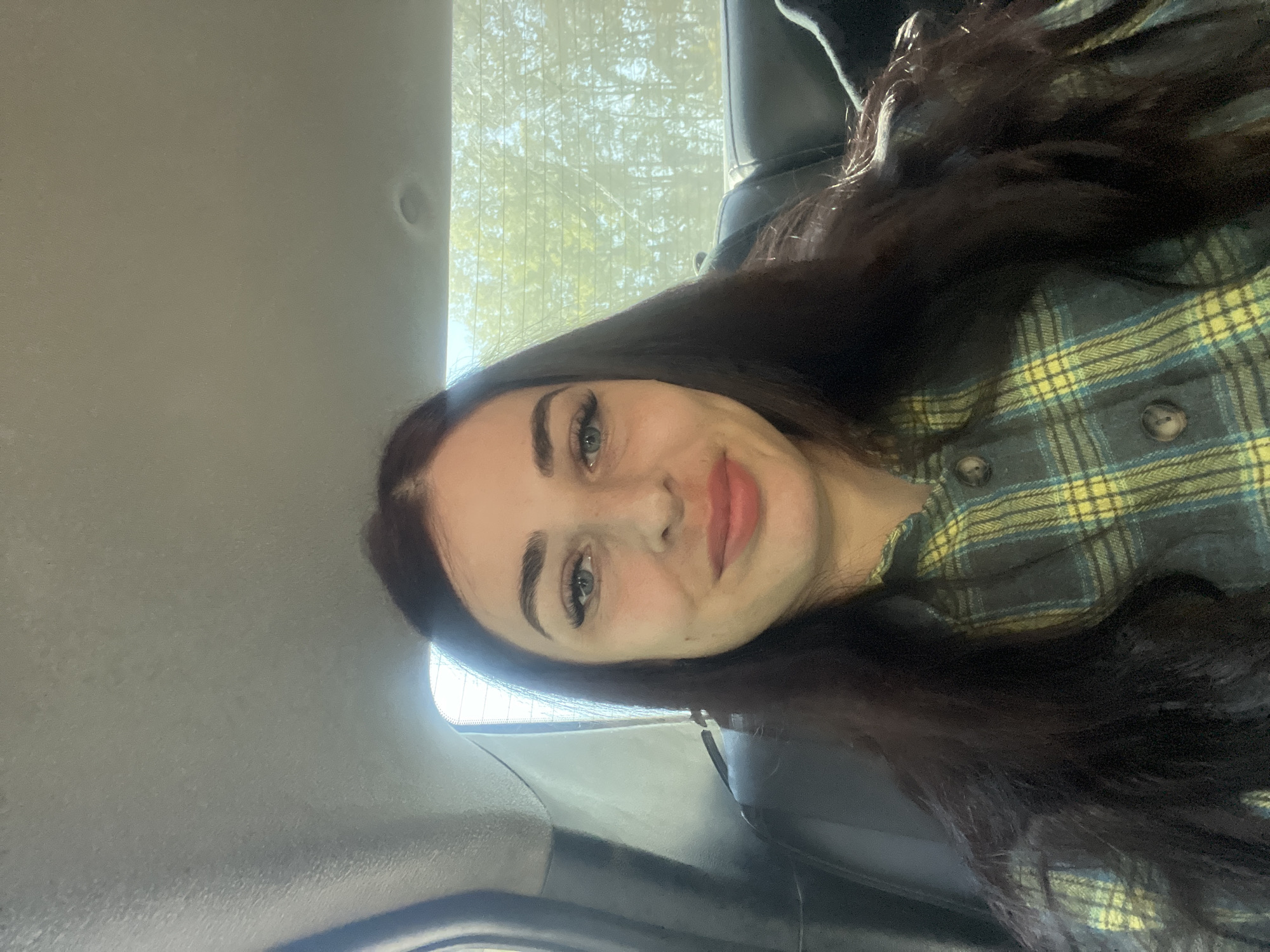A selfie of a woman with long, dark hair, blue eyes, and fair skin, wearing a yellow and green plaid shirt. She is inside a vehicle, likely a car, judging by the interior which includes a window showing trees, and a gray ceiling and side panel. The location cannot be verified as Chaikovsky, Russia with the given information.