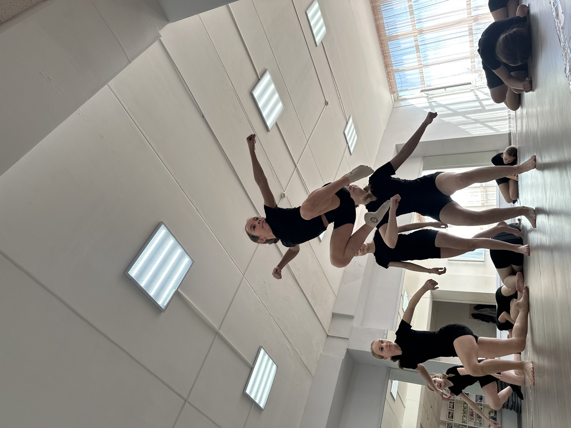 A brightly lit dance studio setting shows a group of young dancers, all dressed in black leotards and shorts, engaged in a rehearsal or practice session. Some dancers are in mid-movement, striking poses that suggest an energetic routine, while others are standing, likely observing or taking a break. The environment includes studio lighting fixtures on the ceiling, a mirrored wall reflecting the dancers and the room's contents, and large windows allowing natural light to filter in. It is unknown if this photo was taken in Votkinsk, Russia.