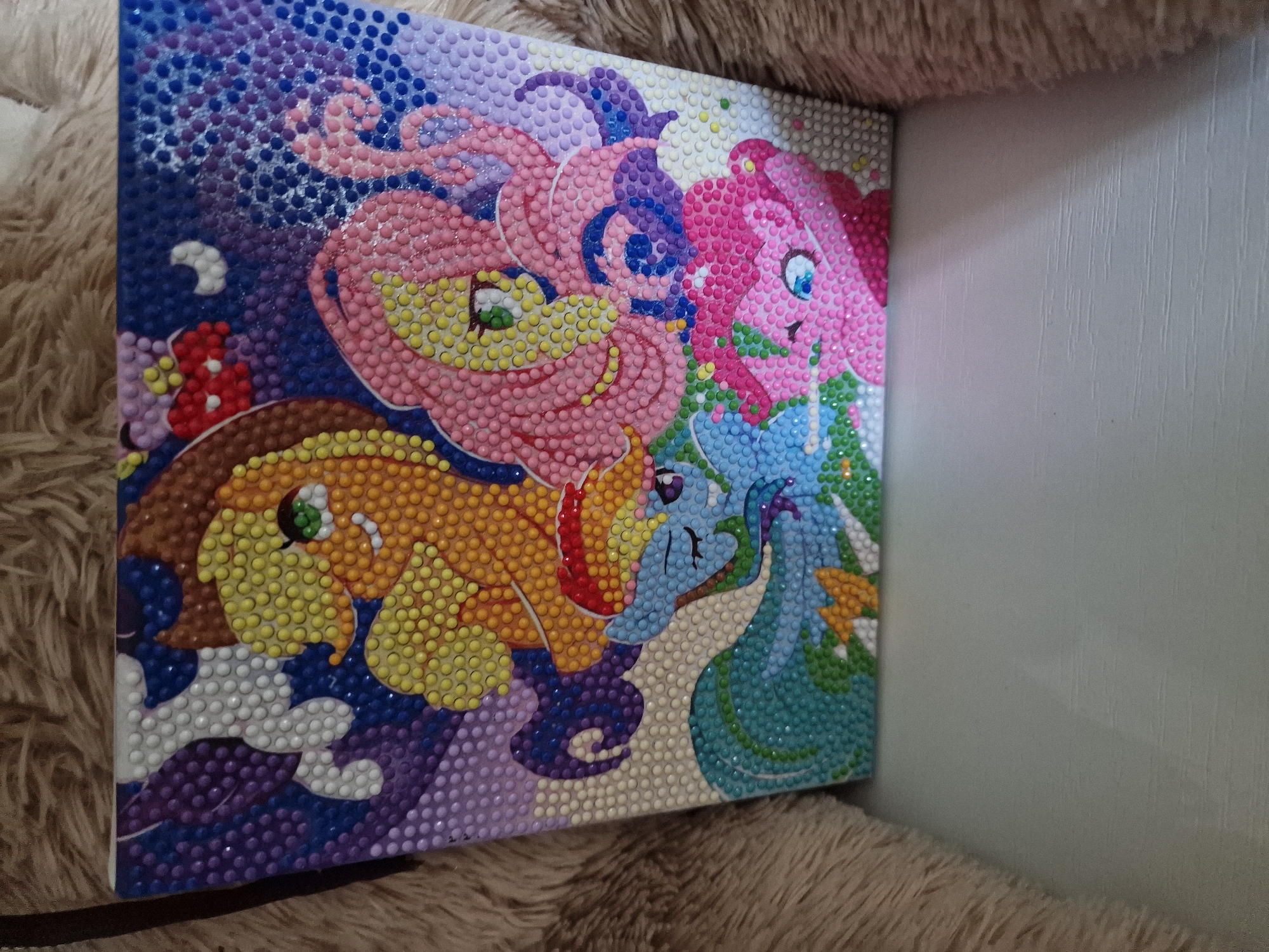 The image shows a diamond painting of characters from My Little Pony. The painting features Applejack, Fluttershy, Rainbow Dash, and Pinkie Pie. The background is a starry night sky with purple and blue hues. The diamond painting is positioned on a white surface, possibly a table or shelf, and is partially obscured by a furry, brown material, likely a blanket or rug. The image does not contain any information about the city of Kursavka, Russia, or any forensic details.