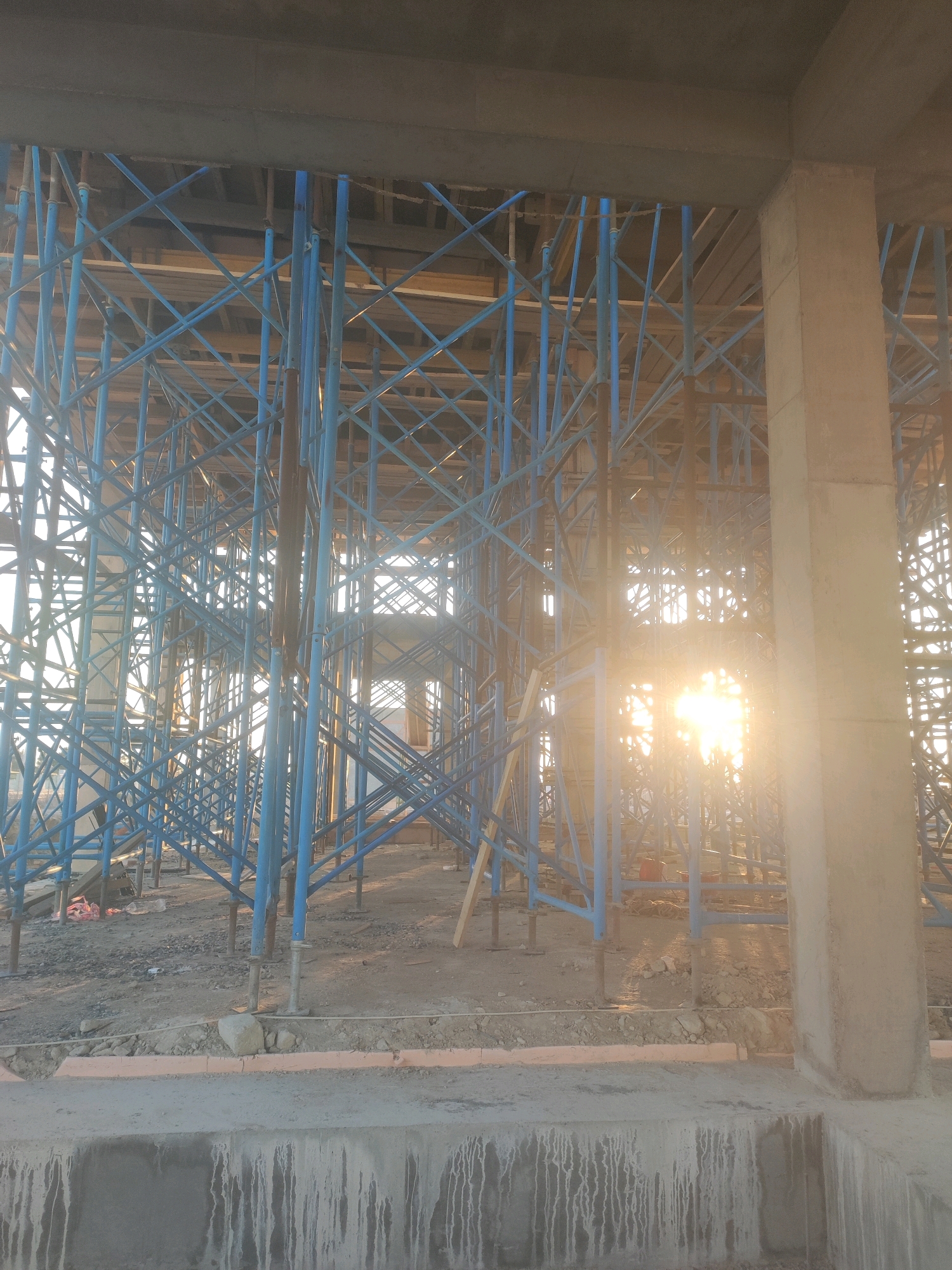 The image depicts a construction site in Khazar Raion, Azerbaijan. The scene is dominated by a complex structure of blue scaffolding, which supports a concrete slab overhead. Sunlight streams through the scaffolding, creating a lens flare effect and illuminating the dusty ground below, which is littered with debris and construction materials. A large concrete pillar is visible on the right side of the frame, adjacent to the scaffolding. The overall impression is one of ongoing construction work in an outdoor environment. There are no people visible in the image.