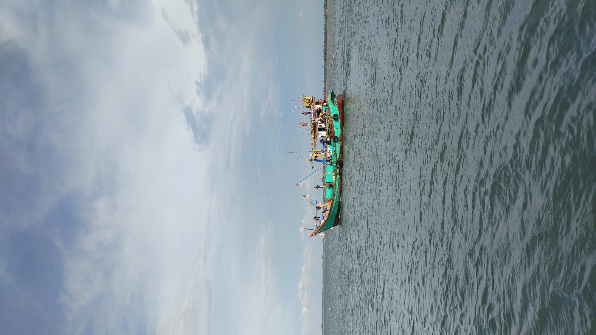 A long, narrow fishing boat with a green hull and red trim is sailing on a body of water. The boat is populated with several individuals, some of whom appear to be actively working. The sky above is cloudy with patches of blue visible. In the distance, a low-lying shoreline with vegetation is visible. The water is slightly choppy, with ripples and small waves. This scene is located in Lhokseumawe, Indonesia.