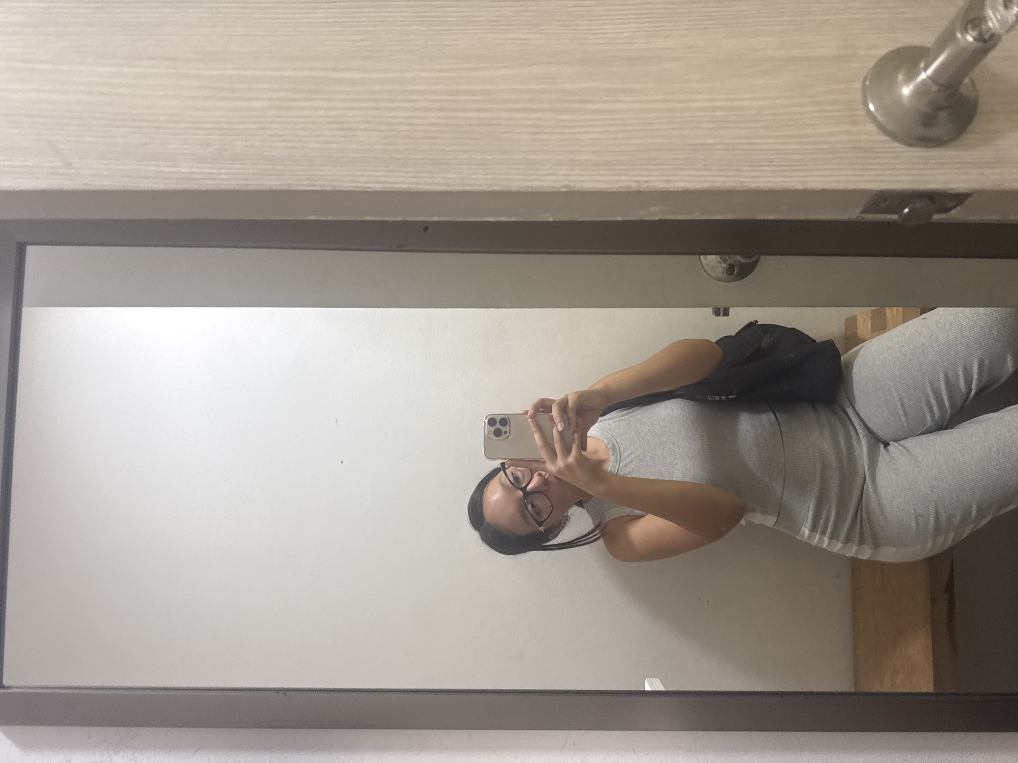 The image displays a mirror selfie taken indoors by a person, likely female. The person, viewed upright in the reflection, has dark hair tied back, wears glasses, and is dressed in a light gray ribbed tank top and matching light gray pants or leggings, which feature a white stripe down the side. A black bag or small backpack is slung over her left shoulder. She is holding a light-colored smartphone (possibly an iPhone) at eye level, capturing her reflection in the full-length mirror.
The background reflected in the mirror behind the person is a plain, light-colored wall. A vertical wooden beam or structure is partially visible to the lower right of her reflection. The mirror is enclosed by a dark frame. Above the mirror frame, a section of a light-colored, wood-grain surface, likely a door or wall panel, is visible, with a metallic door handle positioned in the upper right corner of this surface. The setting appears to be a simple indoor space, possibly a room or hallway. There is no visible text in the image, and while the location context is Puebla, Mexico, the image itself does not contain specific visual cues to indicate this.