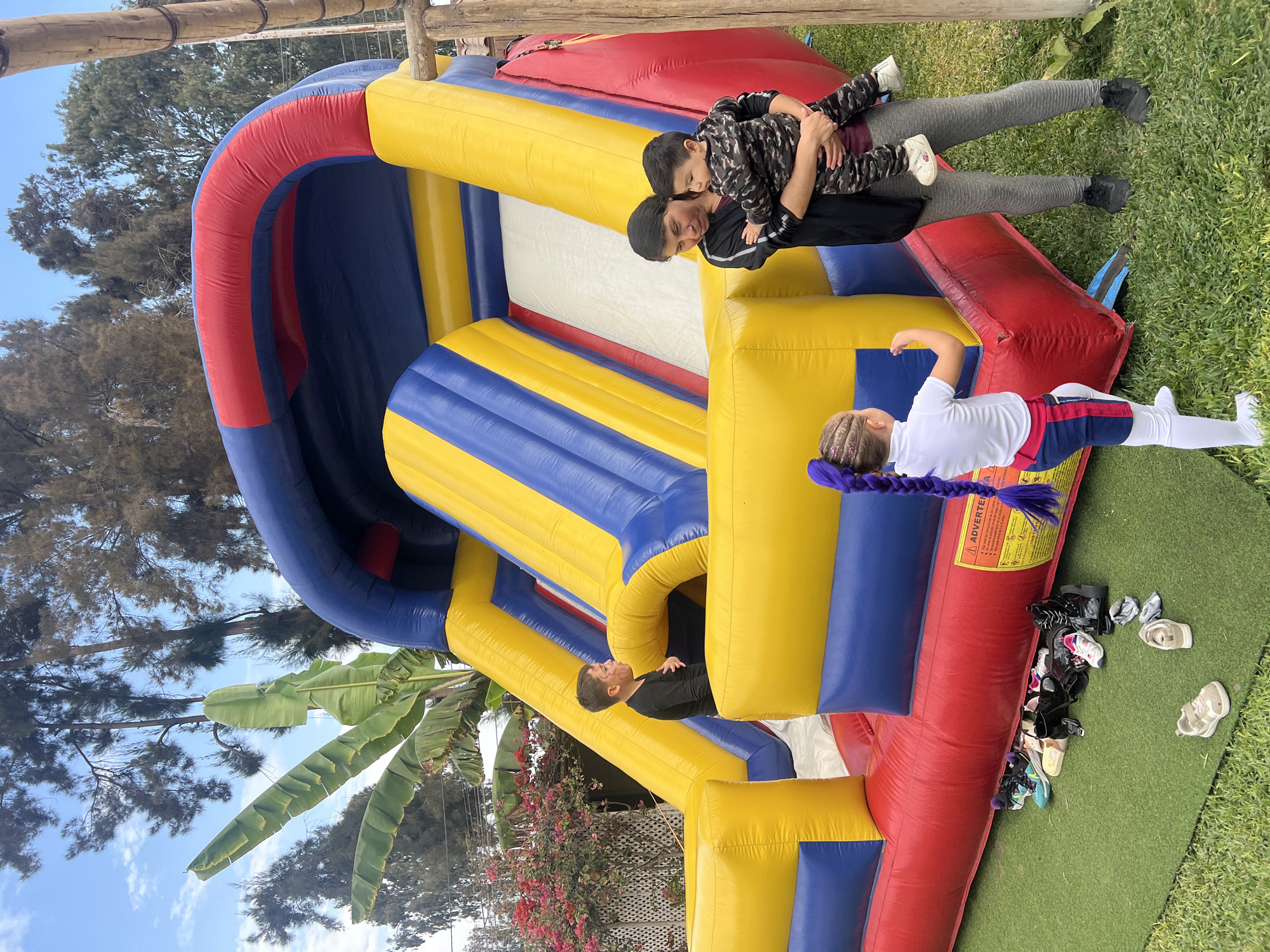 The image shows a large inflatable bouncy castle set up outdoors in what appears to be a residential garden setting. The bouncy castle is predominantly blue, yellow, and red. There are several people interacting with the bouncy castle. One adult is holding a child, another person is inside the bouncy castle. Outside the castle, on the green turf, there are several pairs of shoes. The environment includes foliage, a banana tree and flowers suggesting a warm climate. Based on the foliage, the colors in the image, and the background details, the image was likely taken in Arica, Chile.