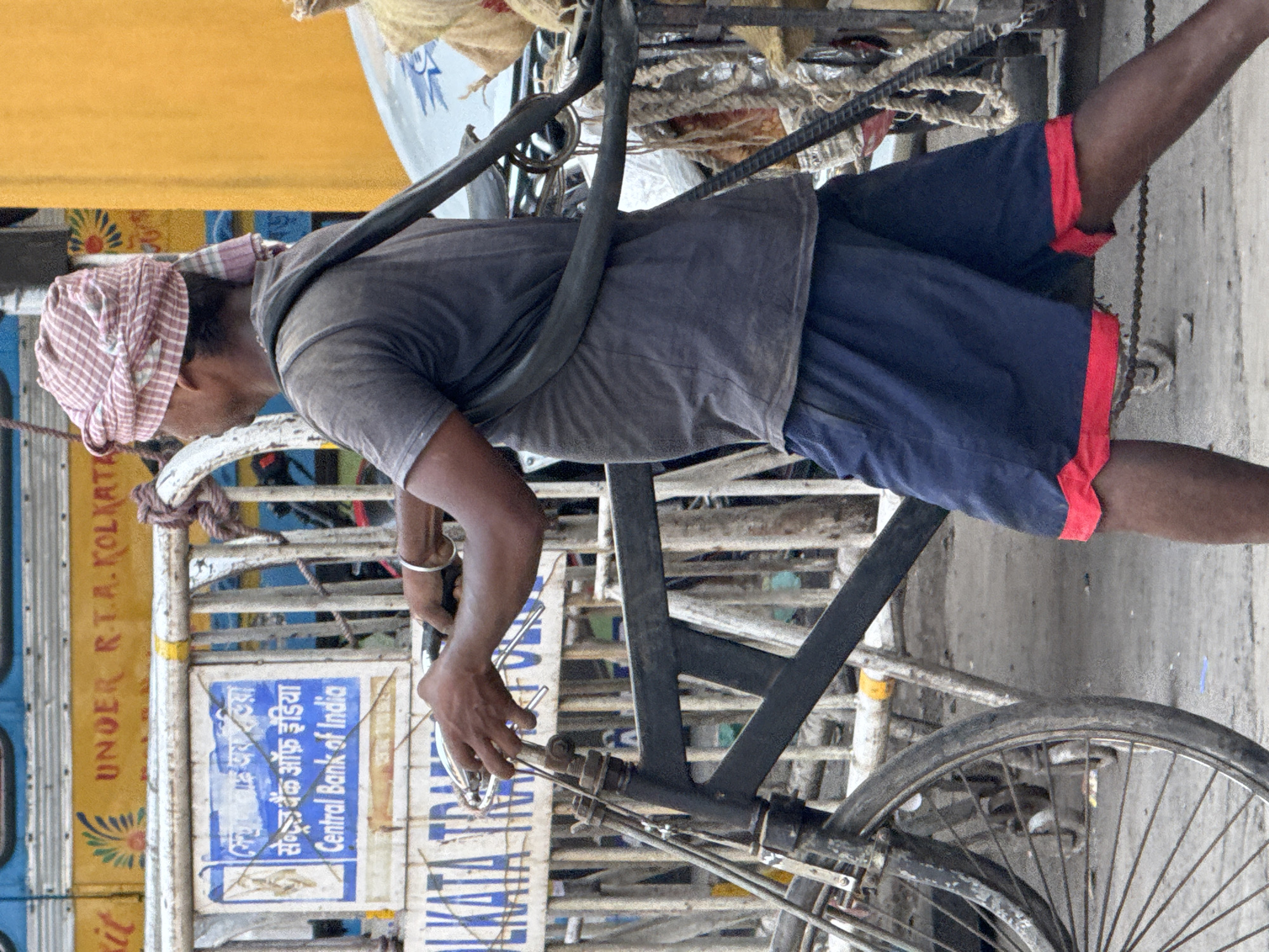 A man with a patterned head covering is pedaling a bicycle rickshaw in Kolkata, India. He is wearing a gray t-shirt and blue shorts with red trim. The rickshaw is loaded with materials, including sacks and ropes. A sign that reads "Central Bank of India" is visible on the side of the rickshaw. The surroundings appear to be an urban street setting.