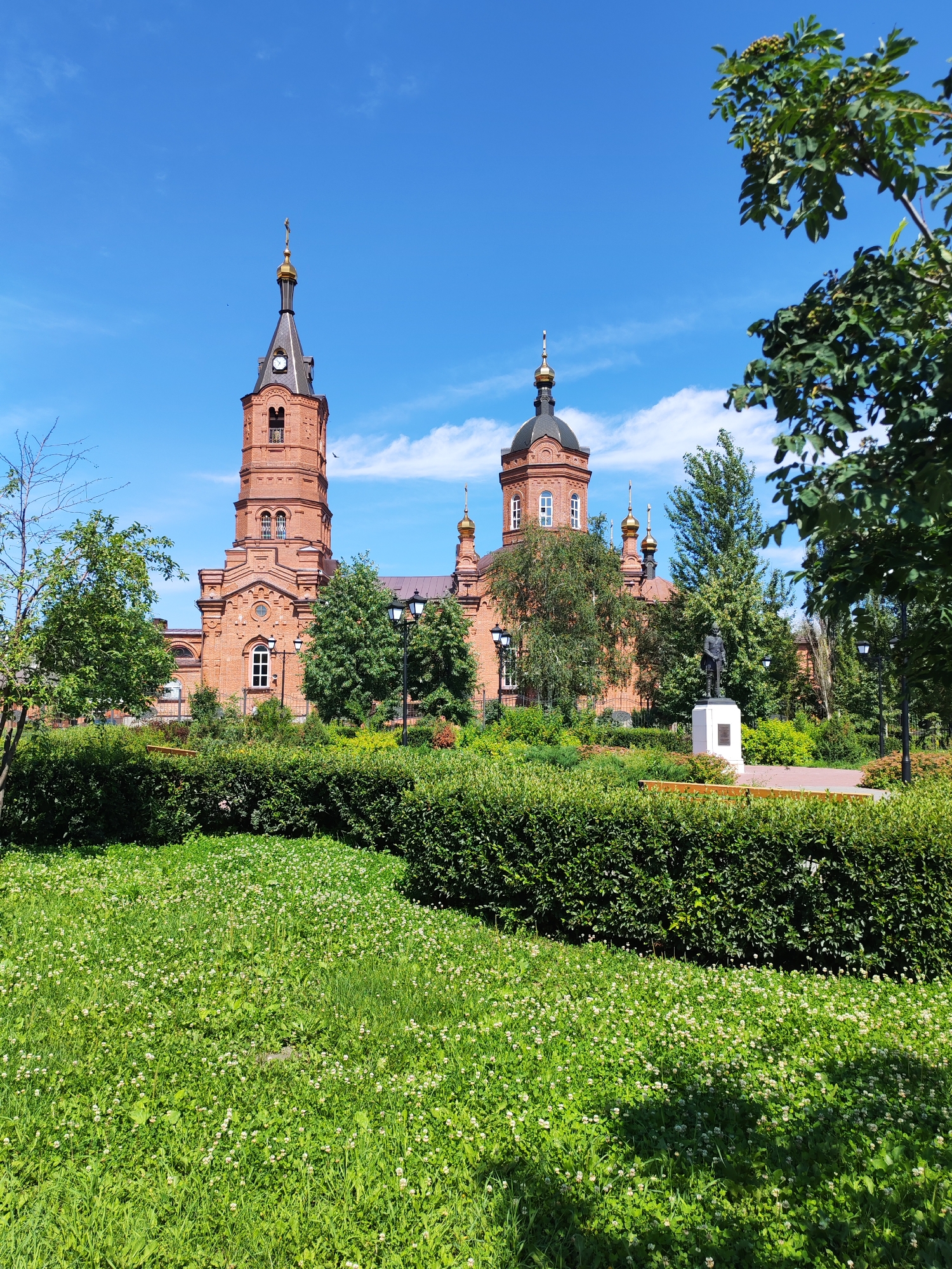 An exterior, eye-level, full shot captures a large, red brick church with multiple domes and a tall bell tower, set against a bright blue sky with scattered white clouds. In the foreground, a vibrant green lawn is dotted with small white flowers, bordered by a neatly trimmed green hedge. Behind the hedge, a paved pathway leads to a statue of a man standing on a pedestal. The scene is illuminated by sunlight, casting shadows. Lush green trees and bushes surround the church and the garden area. The time on the clock tower appears to be approximately 10:30.