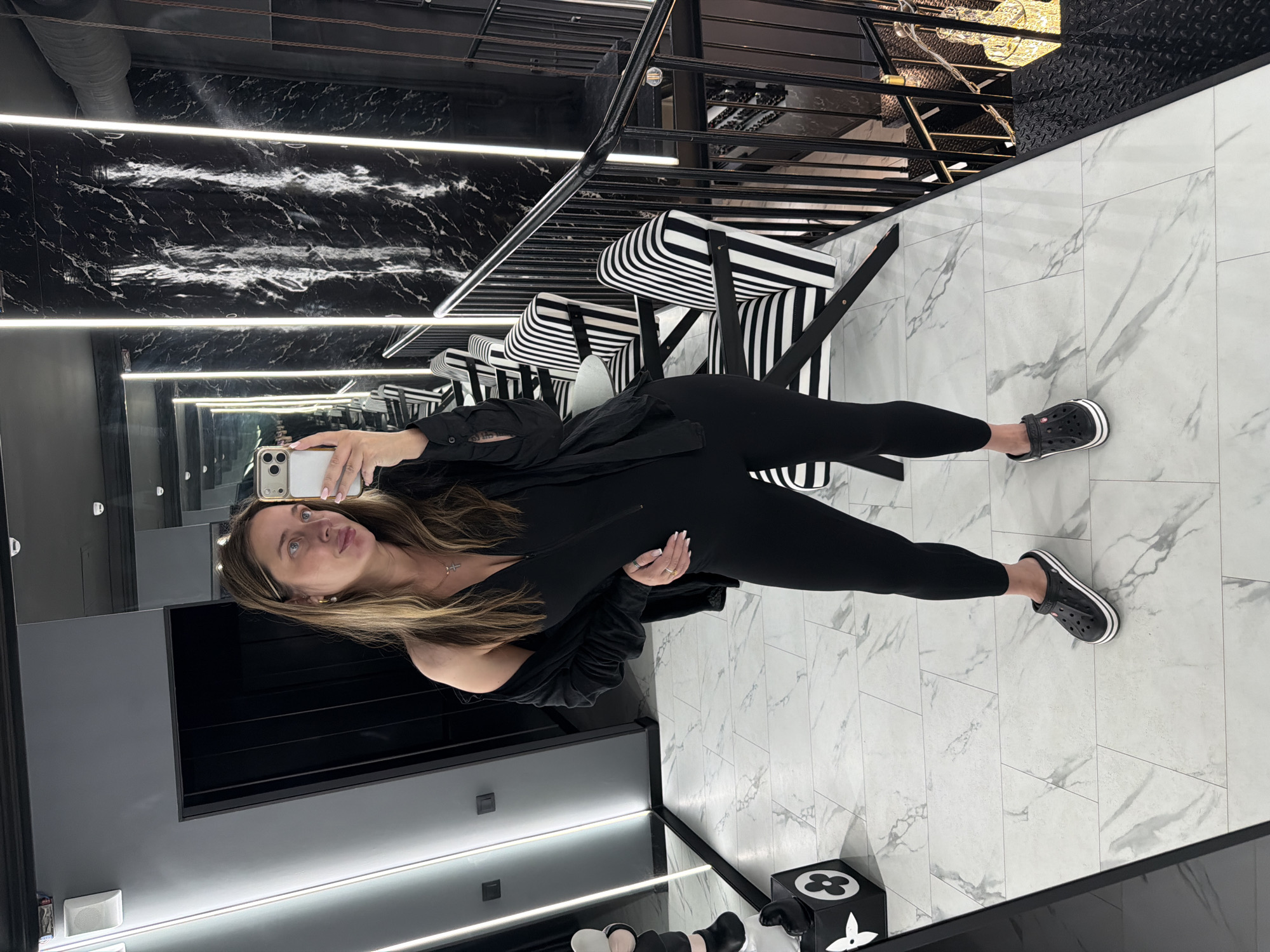 A woman is taking a full-body mirror selfie in what appears to be a modern, upscale retail or commercial space. She is wearing a black, form-fitting jumpsuit with a zipper in the front and black Crocs with white stripes. A black open shirt or jacket is draped over her shoulders. She is wearing a silver cross necklace and a ring on her left hand.
The environment features a black marble-patterned wall with vertical LED strip lighting. To the right, there is a black metal staircase with a black railing and chairs with black and white striped upholstery. The floor is tiled with a white marble pattern. On the left side of the image, there's a mirrored wall reflecting more of the space, and a black cube with a white floral design, possibly a decorative item or a Louis Vuitton inspired piece. The location is the city of Ukromnovske rural settlement, Ukraine.