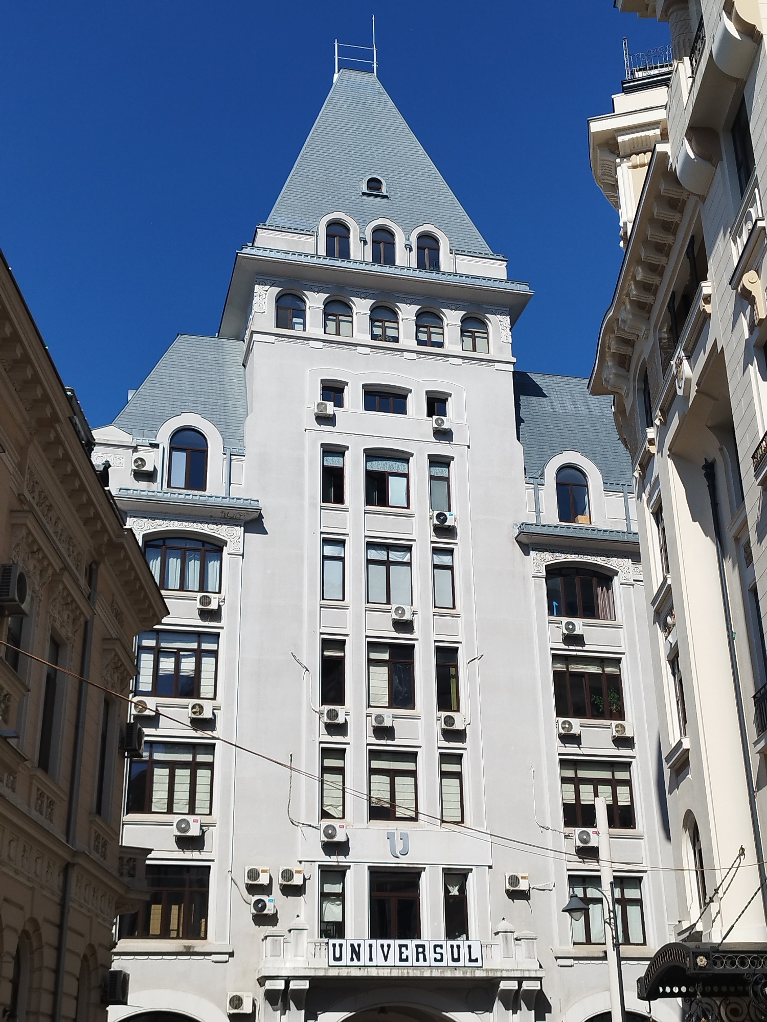 This is a low-angle, wide shot of a grand, light-colored building with a steep, gray, mansard roof, set against a clear blue sky. The building, identified by the signage "UNIVERSUL" at its base, is the Universul Palace, a historical monument located in Bucharest, Romania. It appears to be a multi-story office or residential building, characterized by numerous windows, many of which have dark frames and sheer curtains visible. Several air conditioning units are mounted on the exterior walls, interspersed between the windows on various levels.
The building's architecture is ornate, with decorative elements above some windows and along the roofline. The main structure is flanked by other buildings on either side, one on the left with a similar light-colored facade and ornate detailing, and another on the right which is a lighter, creamy color and appears to be in a classical style with prominent cornices and decorative columns. The perspective suggests the viewer is standing on a street, looking up at the buildings. There are no people visible in the image. The overall scene conveys an urban environment with well-preserved historical architecture.