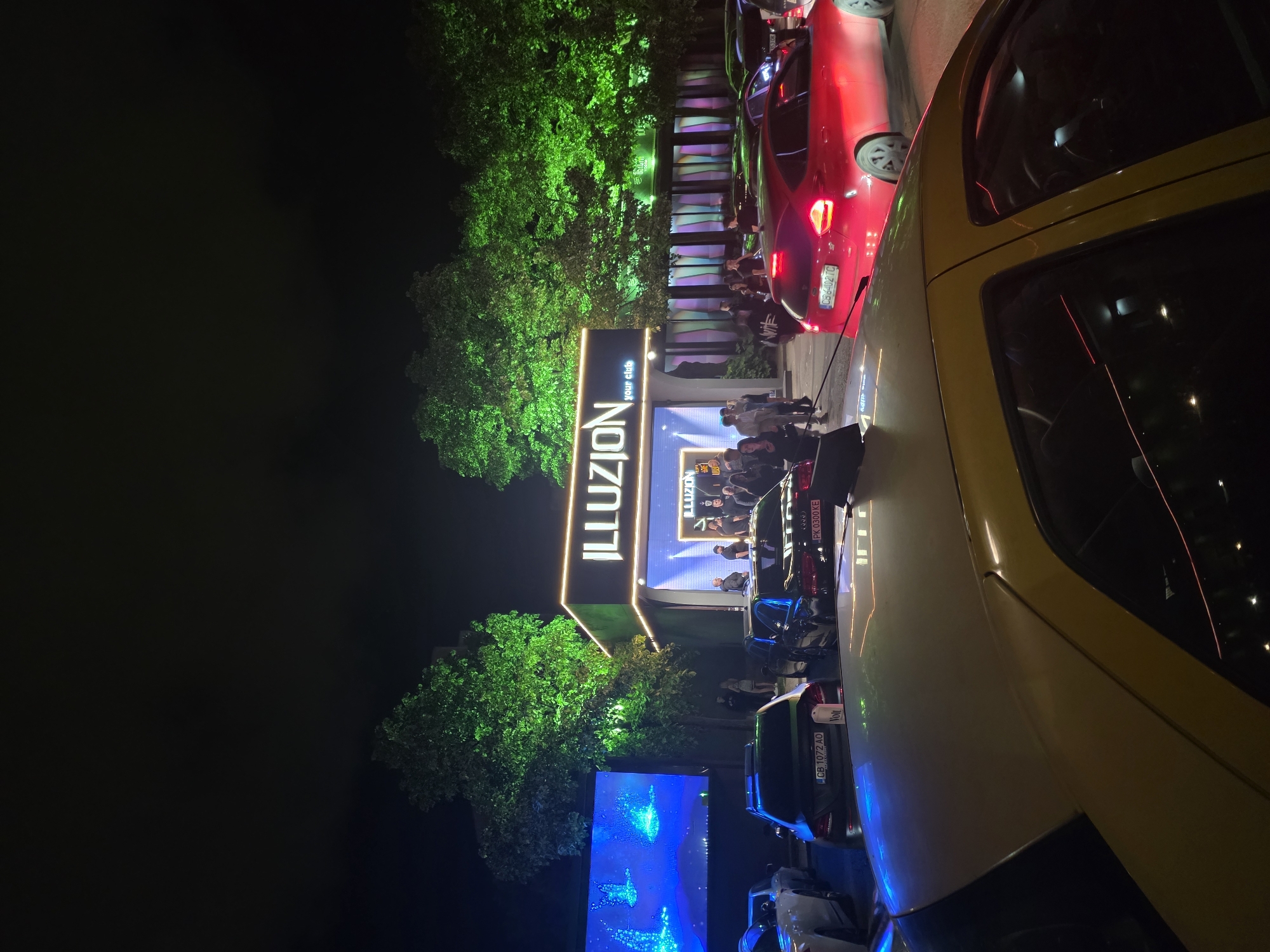 The image depicts an outdoor event at night, possibly a club opening or promotion, identified by the large illuminated sign "ILLUZION your club" in Sofia, Bulgaria. The sign is surrounded by greenery, likely trees illuminated with green lighting. People are gathered near the entrance, suggesting an ongoing event. Several cars are parked along the road or entrance area, with a yellow vehicle prominently in the foreground and a red one further back. The scene is dark, with the event sign, vehicle lights, and illuminated trees providing the main sources of light. A smaller illuminated screen displaying blue graphics is also visible.