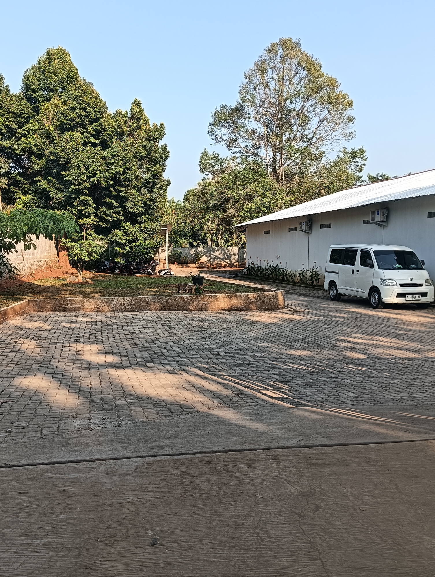 In Cluwak, Indonesia, a white minivan is parked on a paved surface in front of a long, white building with a metal roof. Two outdoor air conditioning units are mounted on the building's exterior. The building is surrounded by vegetation, including trees and low-lying plants. To the left of the building, a grassy area is bordered by a stone wall and features several trees. Further left, a small sapling grows near a larger tree. In the distance, beyond the grassy area, several motorcycles are parked. The ground in the foreground is a light-colored concrete slab, and behind it, a patterned paved area displays dappled shadows from overhead trees. The sky is clear and blue.