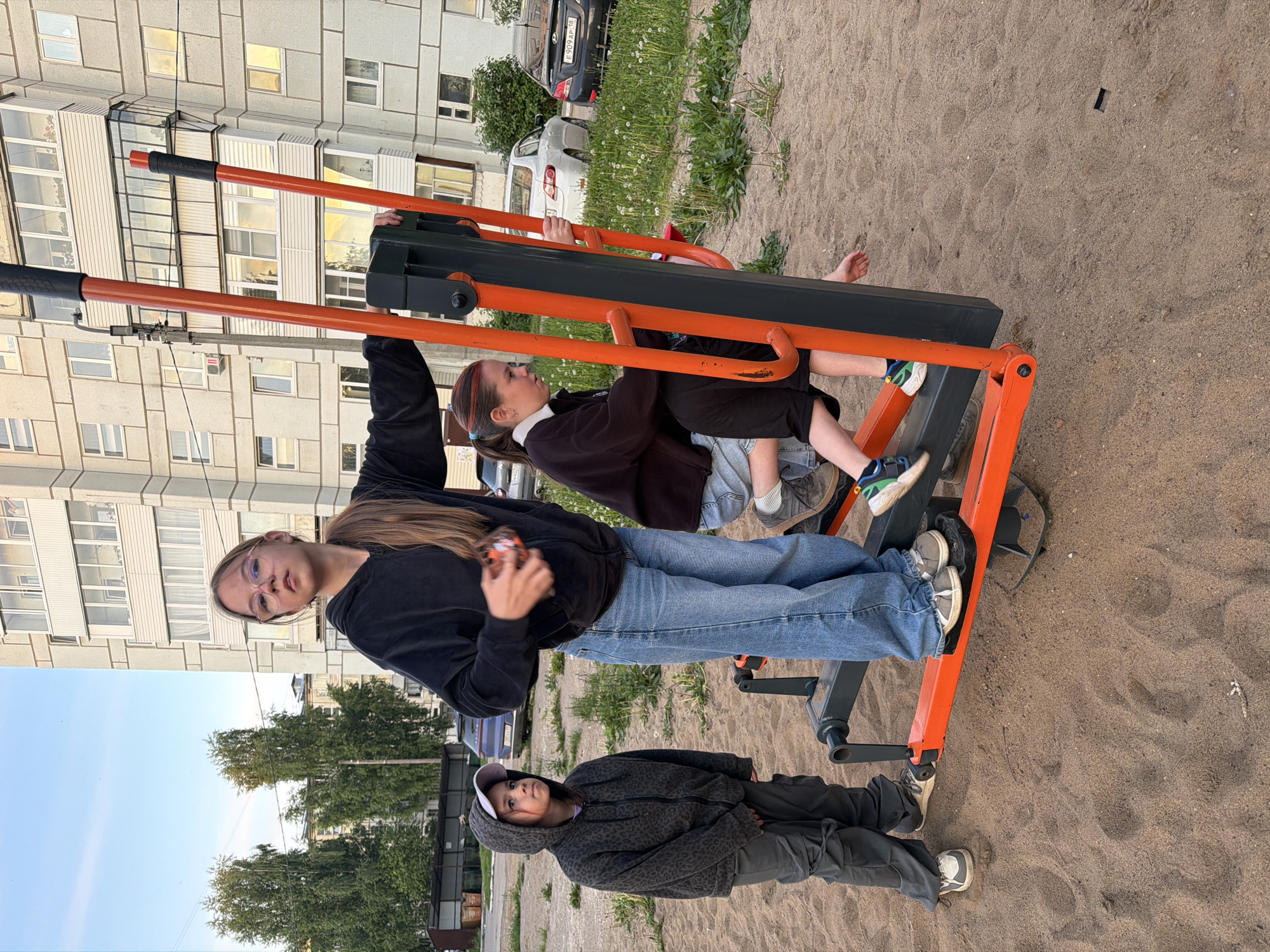 The image depicts three young people engaged in an outdoor scene in what appears to be a residential area of Chaikovsky, Russia. The setting is a sandy patch with an orange and black outdoor exercise machine.
On the left, a girl with long brown hair, wearing glasses, a black hoodie, and blue jeans, stands holding a handle of the exercise apparatus, looking towards the camera. To her right, another girl is seated on the machine, with her feet on the pedals, dressed in a dark jacket and light-colored jeans. She has her hair tied back, revealing some red highlights, and is looking to her right. A younger child, wearing a patterned hoodie, dark pants, and a white baseball cap, stands to the right of the machine, observing the others.
In the background, a large, light-colored, multi-story apartment building with numerous windows and balconies rises, reflecting sunlight in some panes. Trees and greenery are visible between the sandy area and the building, as well as beyond the parked cars. Several vehicles are parked behind the group, including a black car with the Russian license plate "E909AP 18," which corresponds to the Udmurt Republic where Chaikovsky is located. The sky above is clear blue, indicating fair weather. The scene appears to be taking place during daylight hours, possibly late afternoon or early evening, and has a casual, relaxed atmosphere.