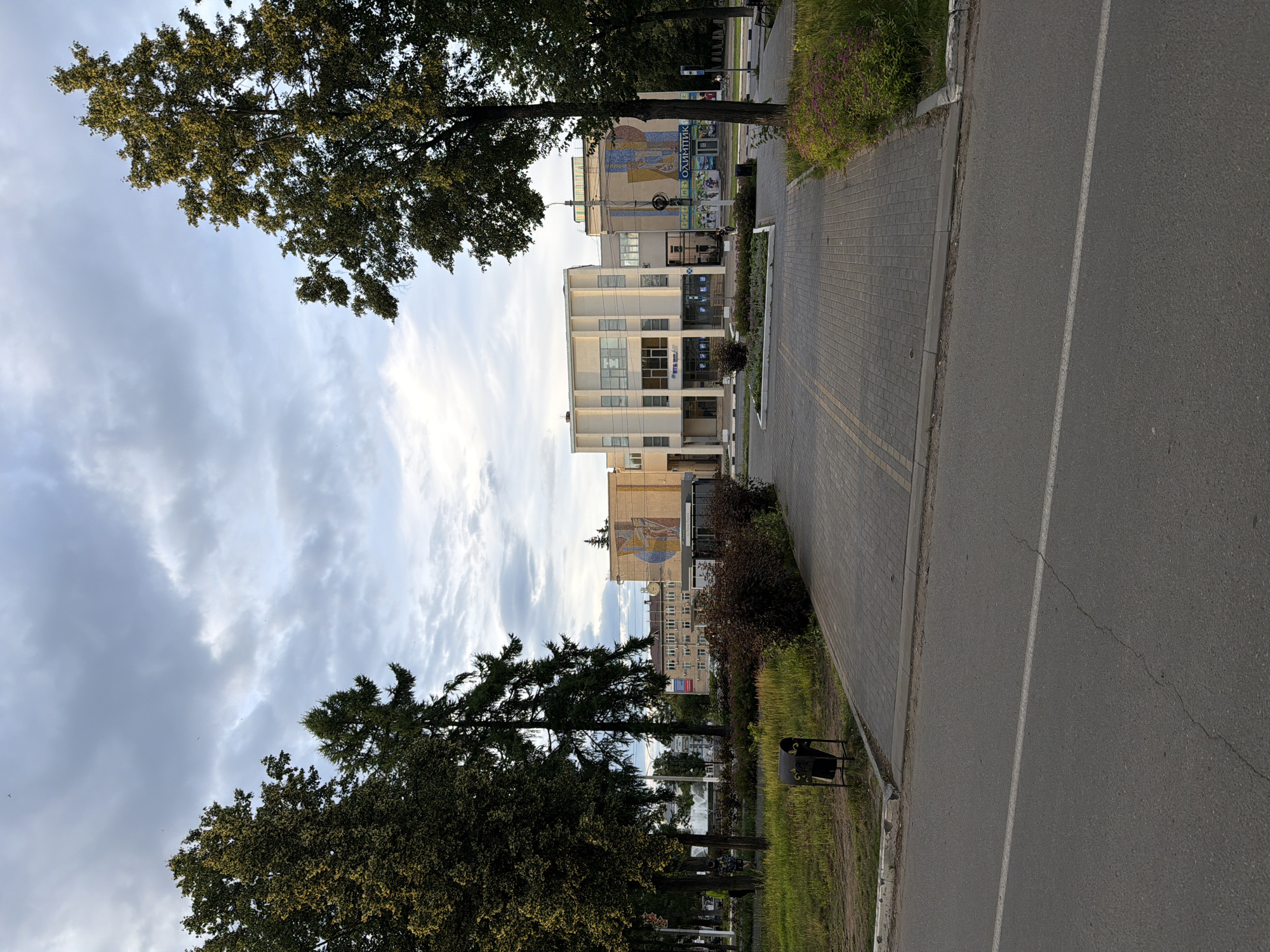 The image depicts a street scene in Chaikovsky, Russia, on an overcast day. The perspective is from the side of a paved road, which has a white line marking the lane. Adjacent to the road is a sidewalk made of grey paving stones, bordered by a concrete curb. To the left of the sidewalk, a strip of green grass is visible, interspersed with trees and bushes. A black trash bin with a decorative pattern is placed on the grass.
In the background, there are several modern buildings with light-colored facades. Some of these buildings have a mosaic mural on their upper floors, one featuring geometric shapes and figures that appear to be part of a larger artwork. There are trees with dense green foliage along the street, framing the buildings. One larger tree with dark green leaves stands prominently in the foreground, its branches reaching towards the sky. The sky is filled with a mix of grey clouds and patches of brighter sky, suggesting diffused sunlight.
The scene appears to be a public space, possibly a city street or a plaza, with a clear emphasis on the architectural elements and the surrounding greenery. No people are clearly visible, and the overall atmosphere is quiet and serene. The time of day is likely late afternoon or early evening, given the quality of light. Visible text on the buildings includes what appears to be "ОЛИМПИАТИК" on a blue banner on one of the buildings.