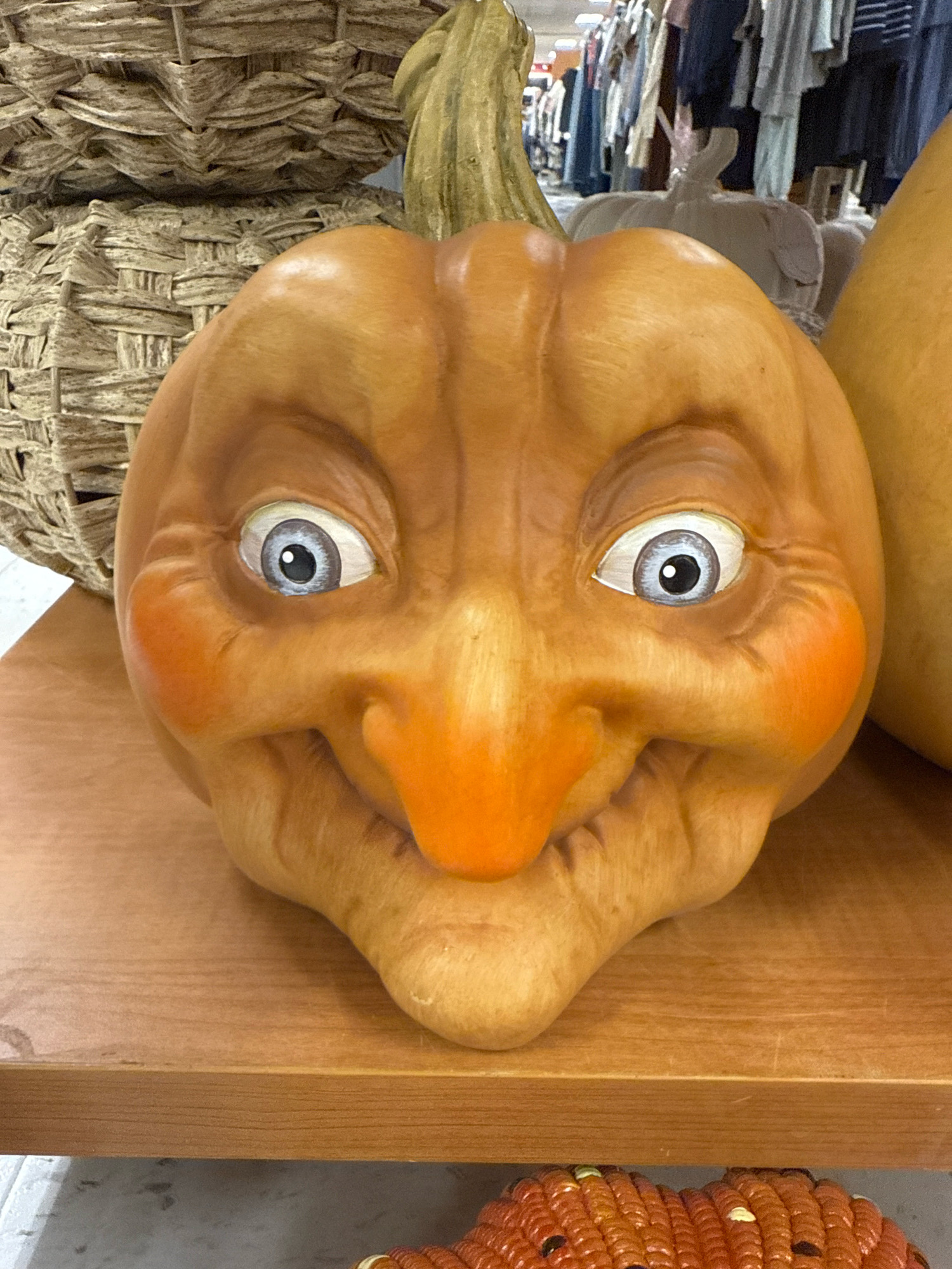 A close-up, eye-level, horizontal shot shows a carved and painted Halloween pumpkin decoration. The pumpkin has a wide, exaggerated grin, rosy cheeks, and large, blue eyes with black pupils. It is situated on a wooden shelf. In the background, a large woven basket is visible to the left, and hanging clothing items are visible to the right. The overall scene suggests a retail environment, possibly a store in Commack, United States of America, that sells seasonal decorations.
