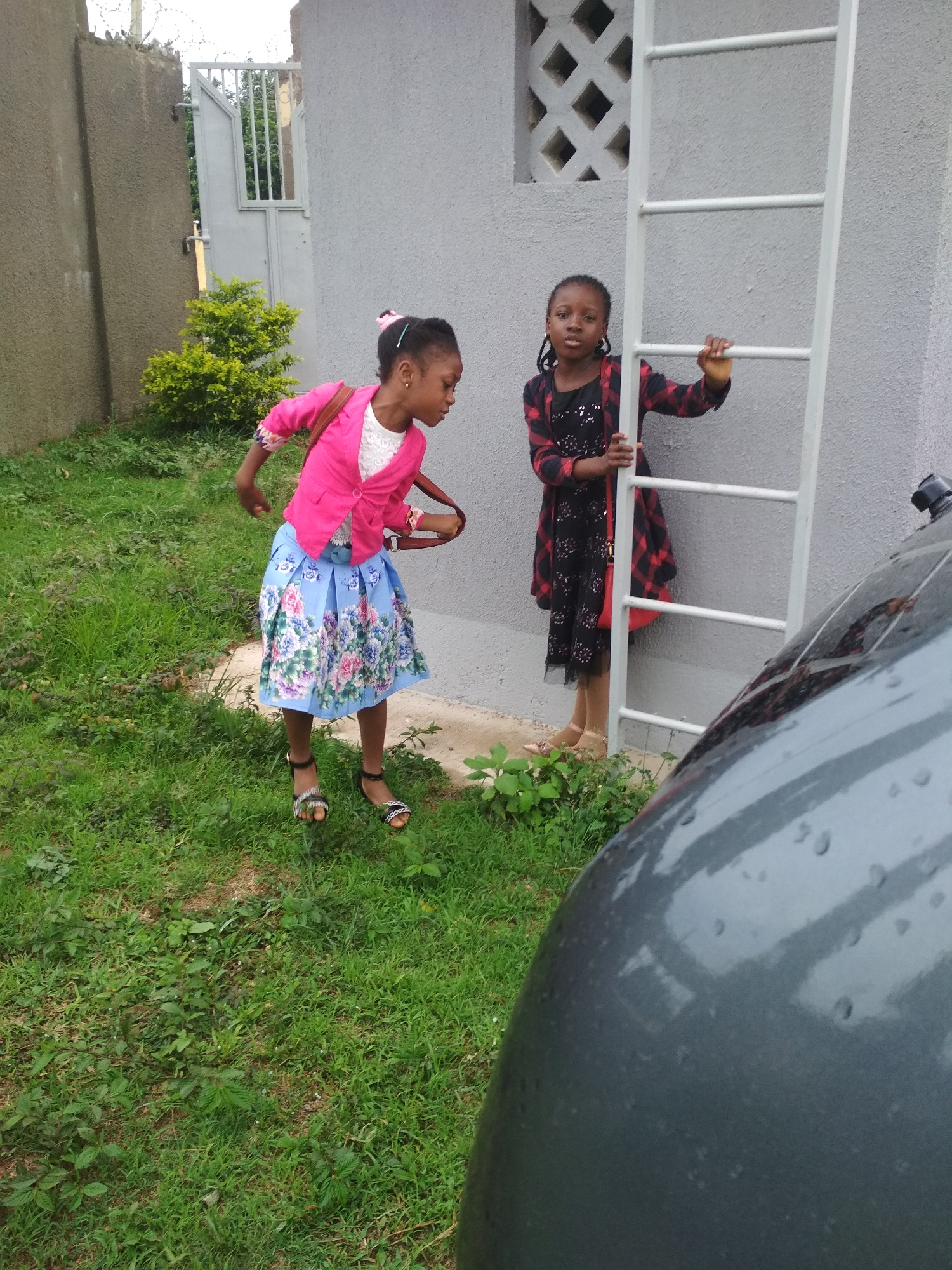 Two young girls are in a grassy outdoor area. One girl, wearing a pink jacket, white top, and floral skirt, stands on the grass with a purse over her shoulder. The other girl stands next to a white ladder, wearing a black and red dress. The setting appears to be a residential area in Jos, Nigeria, with buildings and a gate visible in the background. A portion of a gray vehicle is in the foreground.