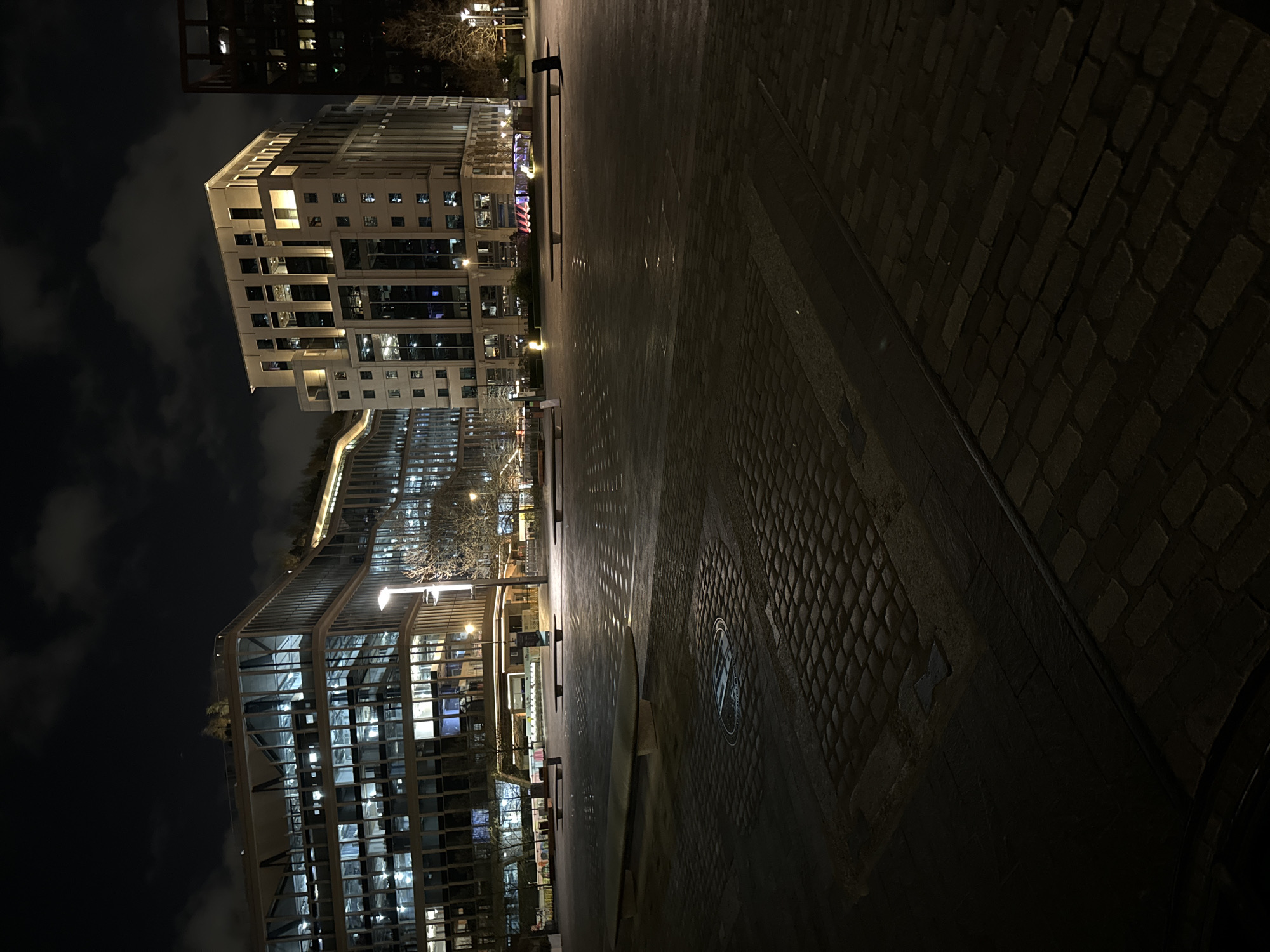 The image captures an outdoor urban setting at night in London, United Kingdom, featuring modern buildings with illuminated windows. The foreground is dominated by a brick-paved area, extending into a smoother, possibly wet, surface that reflects the lights from the buildings. The buildings consist of one that is rectangular and another that is curved with lots of windows. The sky above is dark, with scattered clouds. There are no visible people, actions, or interactions in the frame.