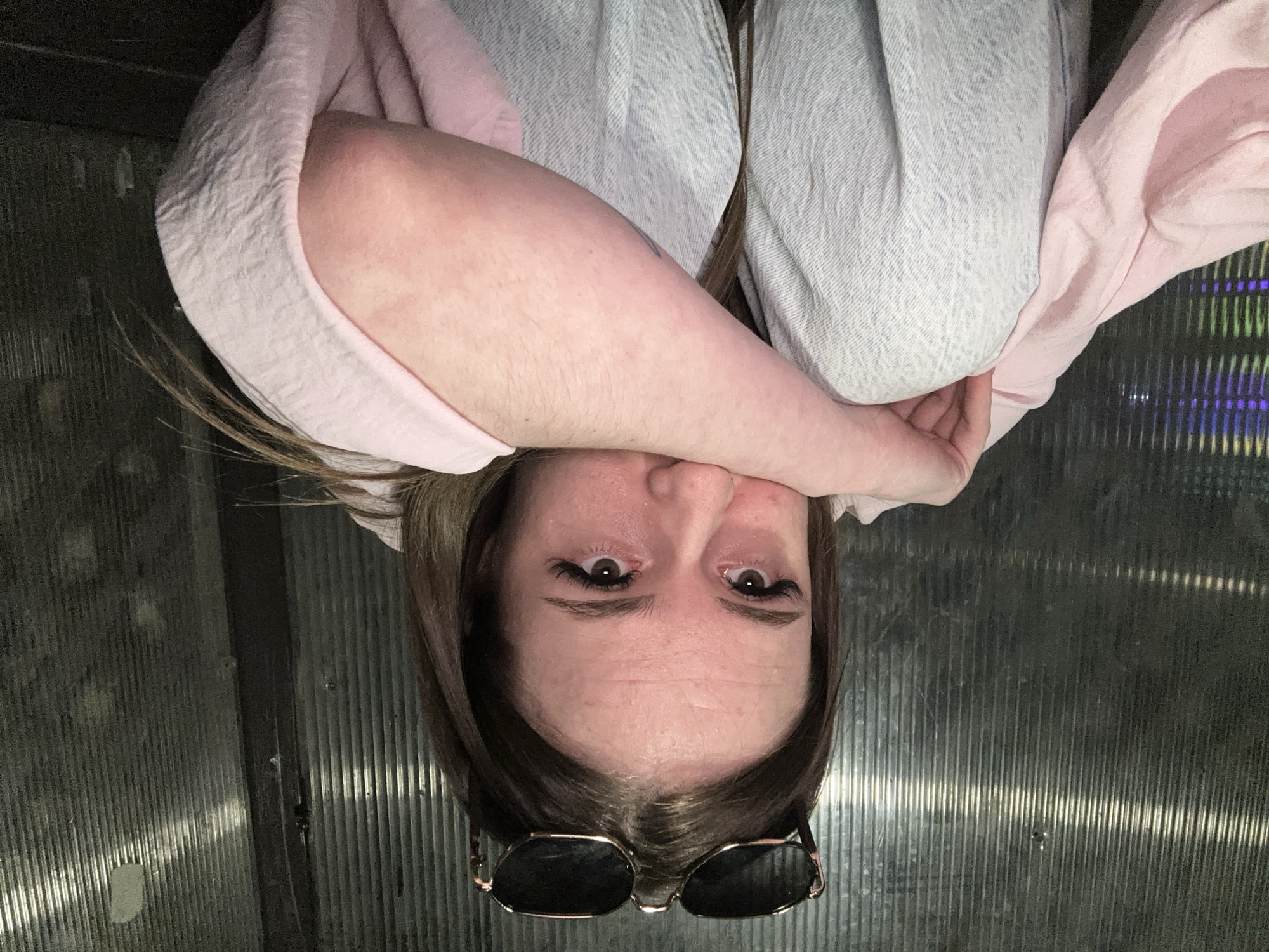 A close-up portrait of a person, likely a young woman, is presented from an inverted perspective (upside down), with her head centered in the frame. Her medium brown hair is parted to the side. Her eyes, adorned with prominent dark eyelashes, gaze directly at the viewer. Faint lines are visible on her forehead. A pair of sunglasses, featuring dark lenses and a metallic (possibly gold or bronze) frame, rests on top of her head.
One of her arms is raised, with the forearm resting across her lower face, partially obscuring her mouth and chin. She is wearing a light pink top or robe. Another garment or blanket, made of a light blue or grey textured fabric with a distinct weave, is also visible, covering parts of her upper body. Her skin tone is fair.
The background consists of a dark, vertically corrugated or ribbed surface, which could be a translucent screen, wall, or window. This surface reflects various light sources, creating bright streaks. On the right side of the image, subtle hints of colored light, such as green and purple, are discernible through the background texture. The lighting appears dim, suggesting an indoor environment or perhaps evening hours. No visible text is present in the image.