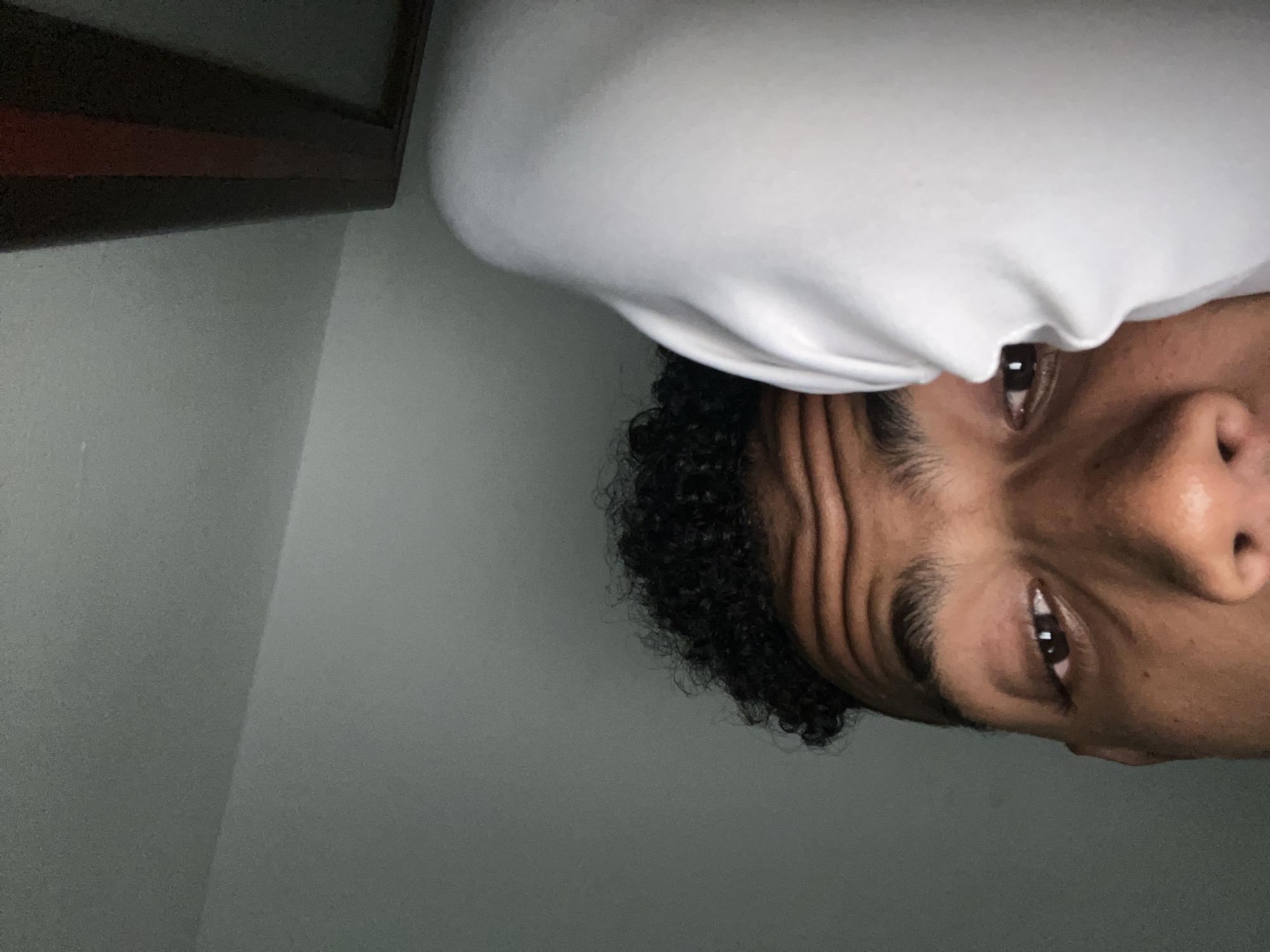 A close-up, vertically oriented photograph shows a person's face partially obscured by a white cloth, likely a head covering. The person has dark curly hair and brown eyes. The background is a gray wall. In the upper left corner, a dark wooden object, possibly furniture, is visible. The photograph appears to be taken indoors. There is no information to determine that this picture was taken in Porlamar, Venezuela.