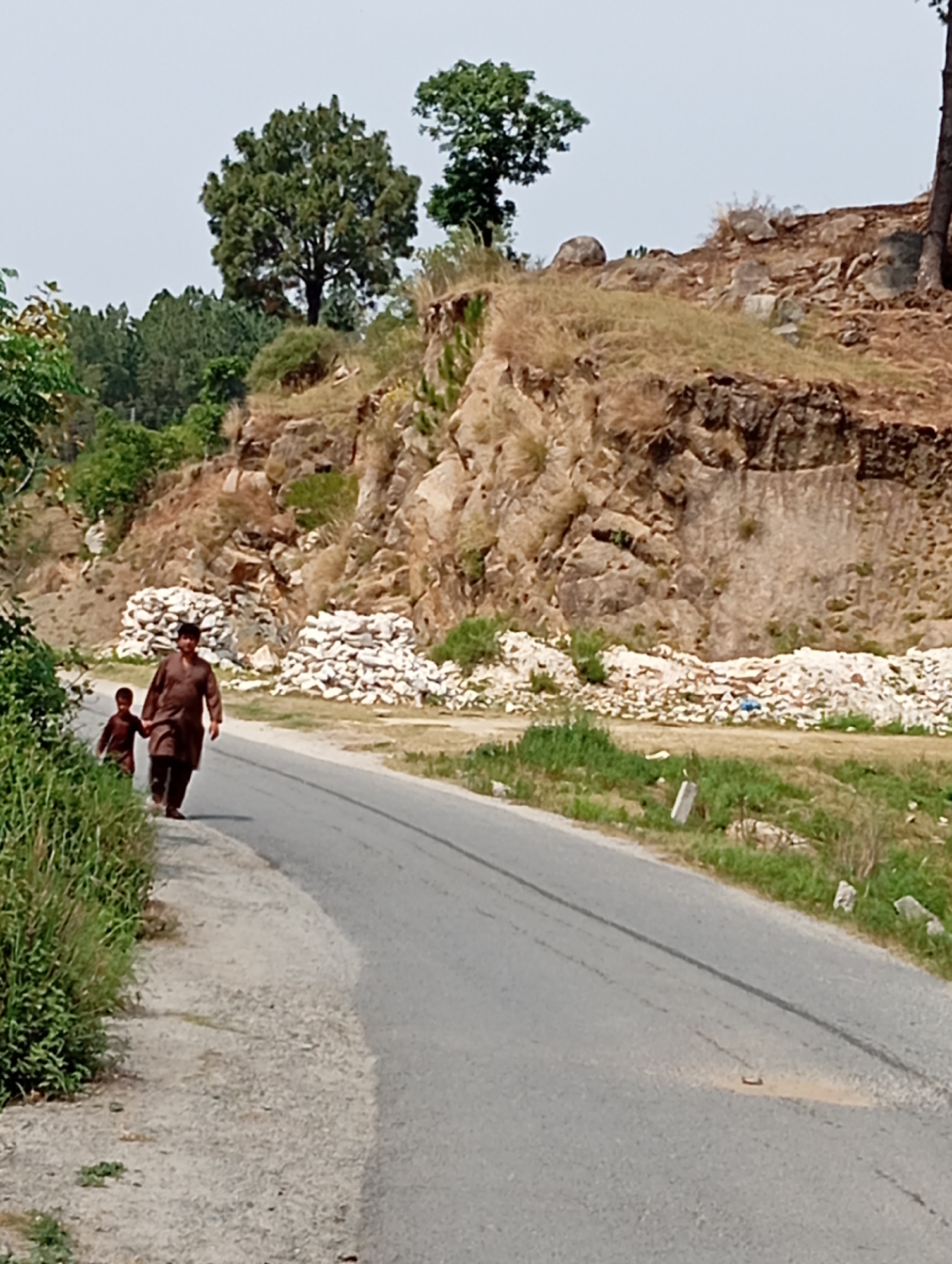 A man and a boy are walking on the side of a paved road. The man is wearing a brown shalwar kameez and the boy is wearing a brown shirt and pants. They are walking away from the camera, towards the right side of the image. The road is empty except for them. The environment is rural, with a rocky hillside on the left side of the road and a steep, rocky embankment on the right. There are some piles of white material, possibly stone or construction debris, at the base of the embankment. Trees and foliage are visible on the hillside and along the road. The sky is a pale blue.