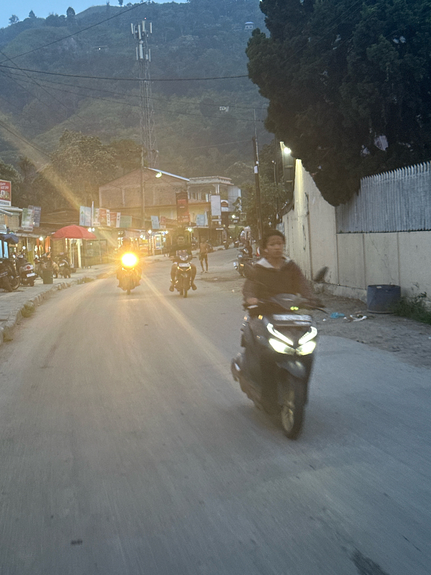 The image depicts a street scene in Parsaoran Ajibata, Indonesia, likely during dusk or early evening. A paved road runs through the frame, with a slight incline. On the right side of the road, a light-colored wall with a grey metal fence is visible. Adjacent to the wall, a blue trash receptacle is present. To the left of the road, a sidewalk or paved area is lined with small shops or stalls, some with red umbrellas.
Several people are on motorcycles, indicating active street life. In the foreground, a person is riding a dark-colored scooter with bright headlights. Further down the road, another motorcycle with a very bright headlight is approaching, obscuring the rider. Other motorcycles are parked along the left side of the street.
In the background, a tall telecommunications tower stands on a lush, green hillside. More buildings are visible on the hillside, as well as trees that cast dark shadows. The overall lighting suggests a transition from day to night. The atmosphere appears to be that of a busy local road.