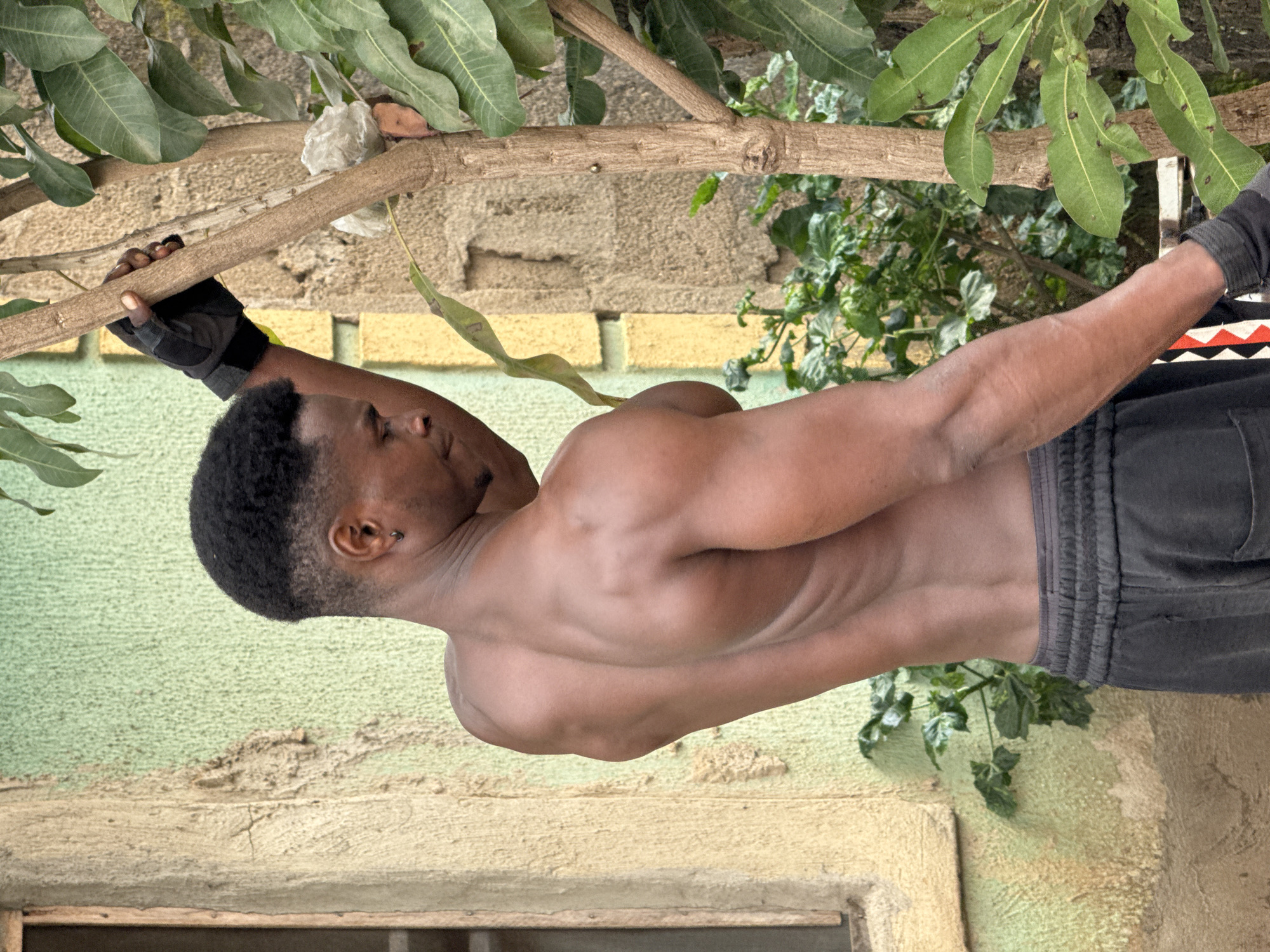 A shirtless man with dark skin, short black hair, and a black earring in his left ear is standing outdoors. He is wearing dark gray sweatpants and black gloves. His left arm is bent, and he is holding onto a tree trunk. He is looking down to his right. The background consists of a textured wall, a window frame, and lush green foliage from trees and plants. The wall appears to be painted a light green and yellow. The scene is likely in Akwanga, Nigeria, given the context of the request.