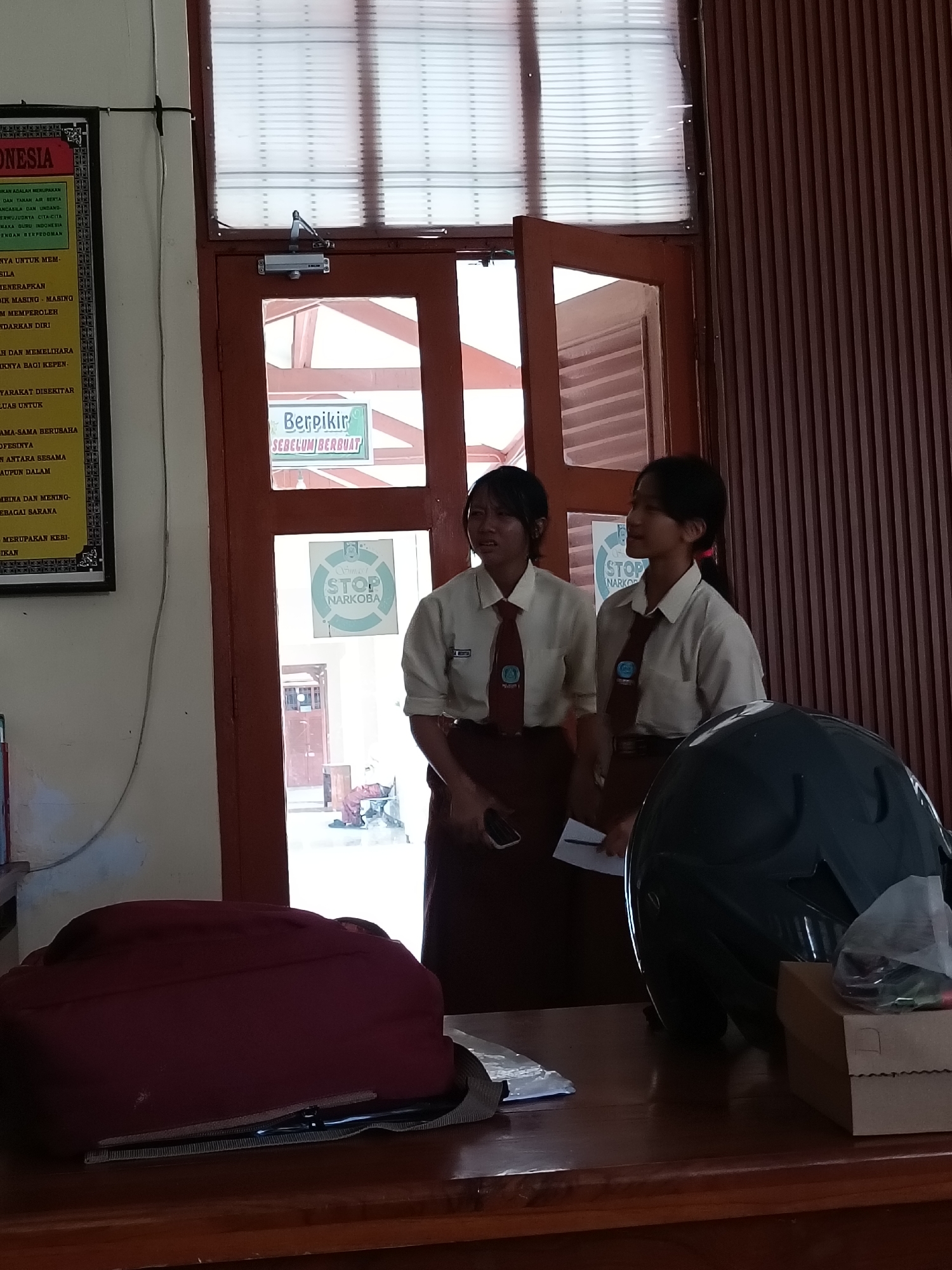 The image captures an indoor scene with two young women in school uniforms standing near a doorway. One woman on the left is wearing a cream-colored shirt and a maroon tie, with her hair pulled back. The other woman on the right is similarly dressed and looking to her left. Behind them, a sign with green lettering on a white background reads "Berpikir SEBELUM BERBUAT" (Think Before Acting). Further down and to the left, another circular sign with a white background and green border has "STOP NARKOBA" (Stop Drugs) written on it in green. The doorway opens to an outdoor area where a person in a red shirt and dark pants is visible. A dark red backpack and a dark green helmet are placed on a wooden table in the foreground. The background features vertical wooden paneling and a window with horizontal blinds. The context provided by the signs suggests a school environment in Indonesia, potentially within Kediri City, Indonesia, given the Indonesian language used on the signs. The overall atmosphere is casual, possibly during a break or after school hours.