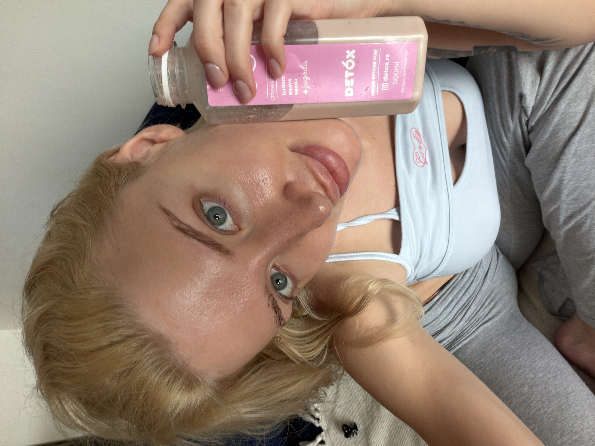 The image shows a young woman with fair skin, blonde hair, and striking blue eyes holding a bottle of "Detox" juice. The juice bottle has a pink label with the ingredients listed as strawberry, almond, agave, vanilla, and strawberry (jagoda). The label also has an Instagram handle "@detox.rs" and a volume of 500ml. She's wearing a light blue tank top with a white sports bra underneath, and grey leggings. The woman is indoors. It is not possible to determine the location in Belgrade, Serbia from the provided image.