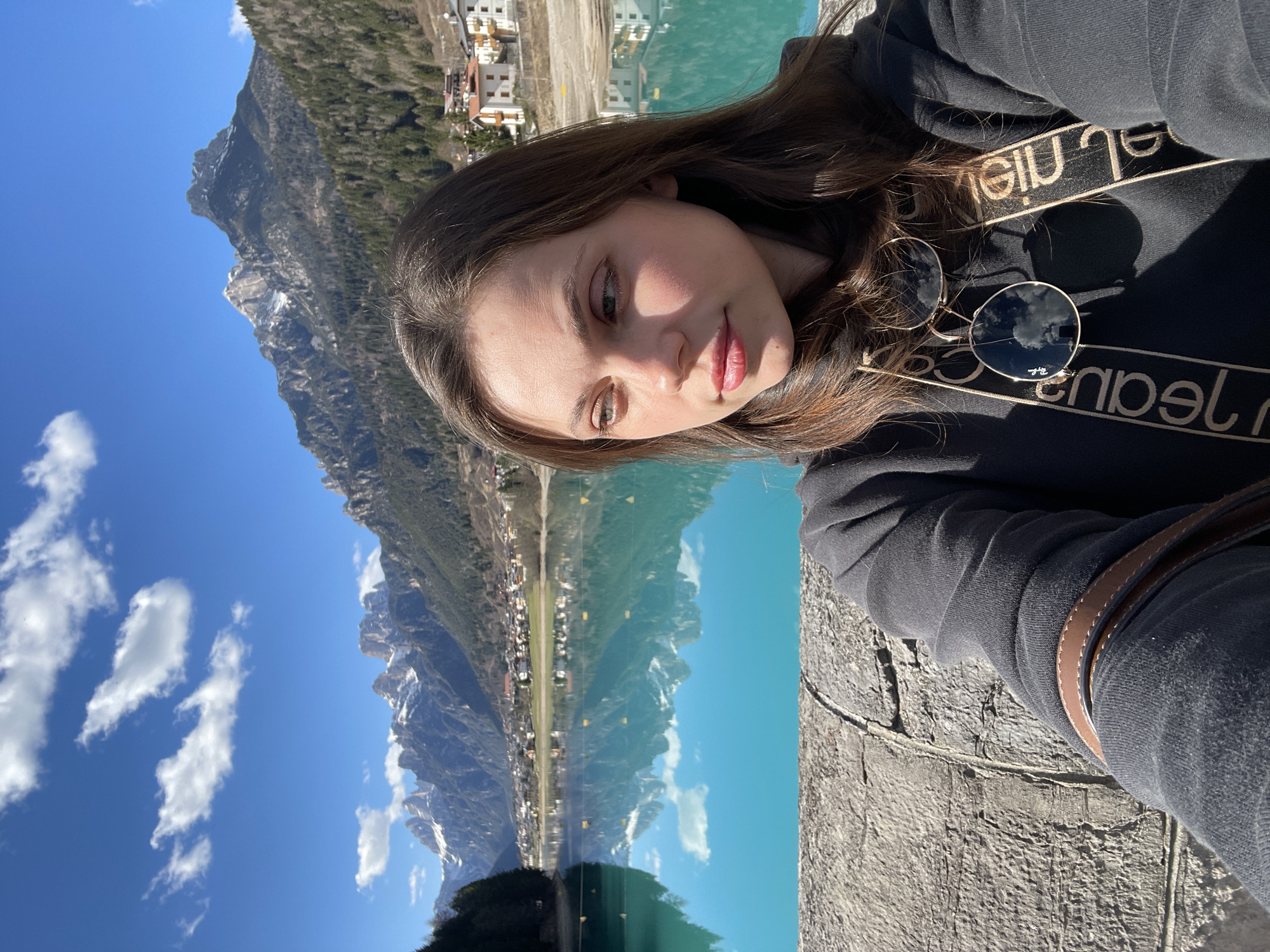A vertically-oriented medium shot features a young woman in the foreground, with a picturesque landscape visible in the background. The woman has long brown hair, fair skin, and is wearing a black sweatshirt with white lettering along the sleeves and a pair of sunglasses resting on the shirt.
The background shows a turquoise-colored lake reflecting the surrounding mountains and a clear blue sky dotted with white clouds. Adjacent to the lake, there is a small town with buildings. The location is Auronzo di Cadore, Italy. The woman appears to be taking a selfie while standing on a stone structure or wall.