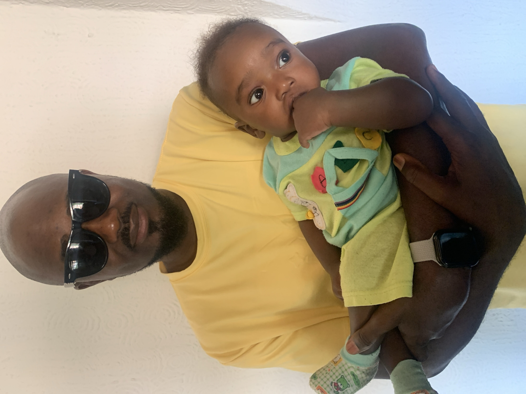 A medium shot of a dark-skinned man holding a baby. The man has a shaved head, a mustache, and is wearing sunglasses and a yellow t-shirt. He also has a watch on his left wrist. The baby has dark skin and curly hair, and is wearing a light green and yellow onesie with cartoon characters on it. The baby is also wearing green and white socks. The baby is looking up with its mouth open with its hand inside. The background is a white wall. The location is Barakin Akawo, Nigeria.