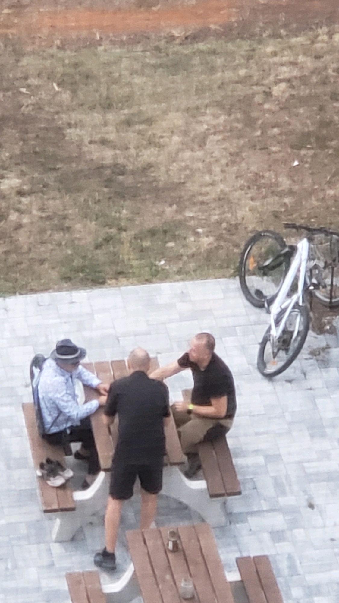 An aerial view shows three adult males gathered around two wooden picnic tables on a paved area in Prešov, Slovakia. In the foreground, a man in a black t-shirt and black shorts stands facing away from the camera, while another man in a black t-shirt and khaki shorts sits on a bench to his right. To the left, a man in a light-colored patterned shirt and a dark hat sits on a bench, his back to the camera. He is also wearing sunglasses. A bicycle with a white frame and black tires is parked to the right of the scene, leaning against something out of frame. The ground beyond the paved area is covered in dry, brown grass. The overall impression is a casual outdoor gathering.