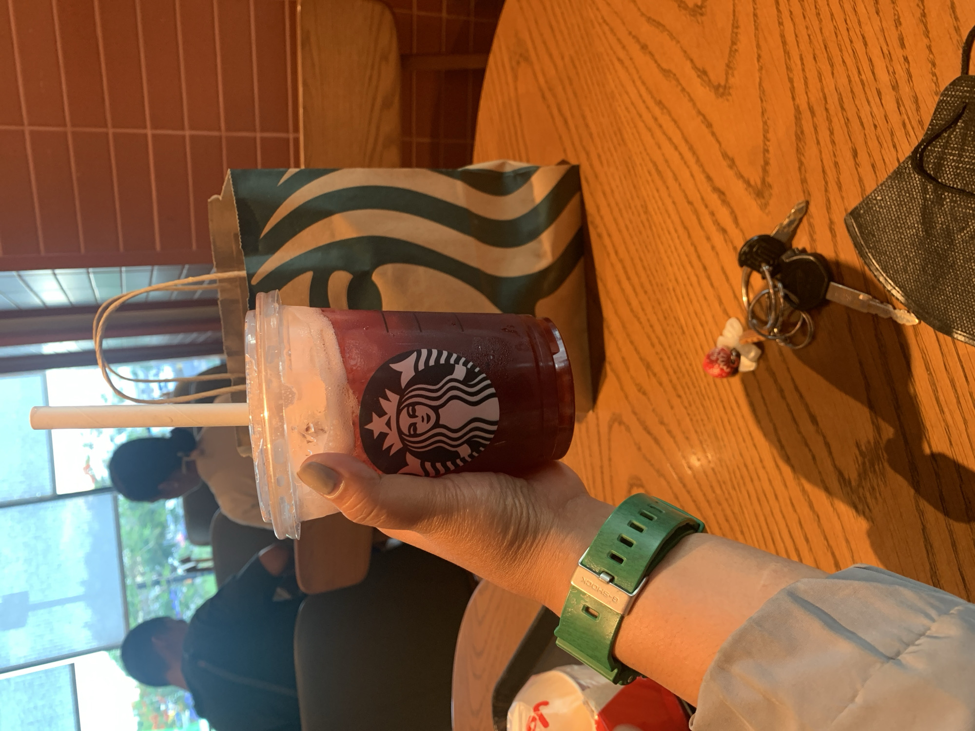 In the city of Angeles, Philippines, a person's hand holds a Starbucks iced beverage with a white paper straw. The person is wearing a green G-Shock watch. A Starbucks paper bag with green stripes is placed on a wooden table. Also on the table are a set of keys, a small red and white decorative item, and a black face mask. In the background, two people are seated near a window with light-colored blinds. The environment appears to be an indoor cafe.