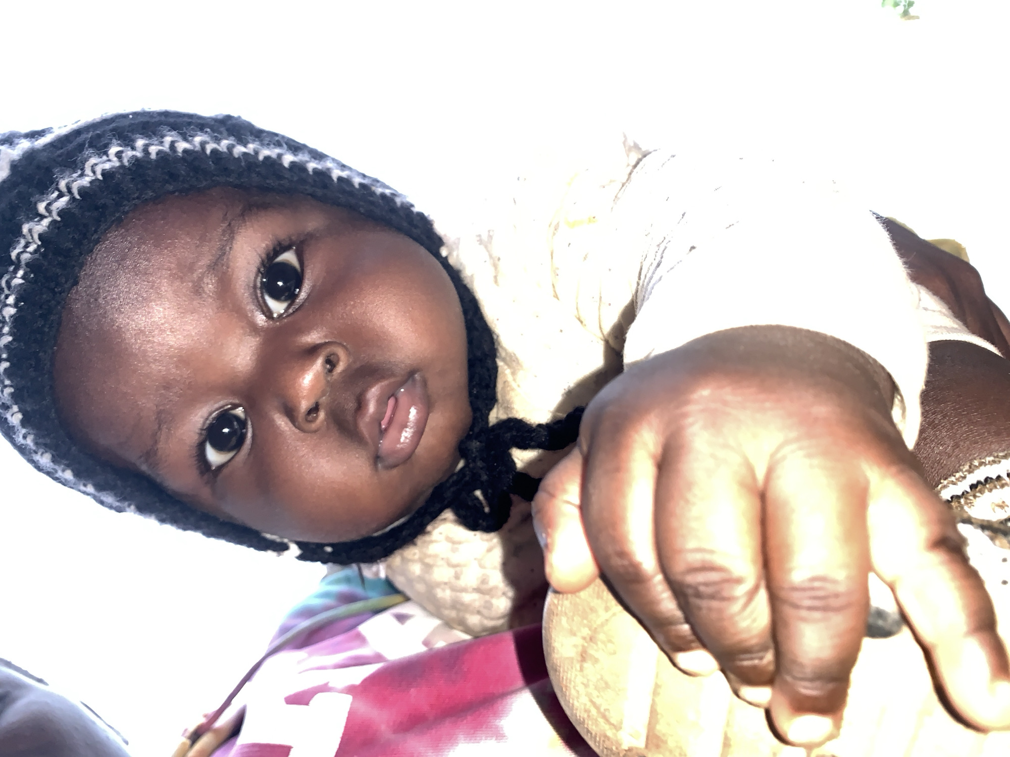A close-up shot shows a dark-skinned baby wearing a knitted hat with black and white stripes around the brim. The hat has black ties under the chin. The baby is wearing a light-colored knitted top. A large hand is resting on a light-colored object, and the baby is lying on a pink and purple patterned surface. The location is presumed to be Akwanga, Nigeria.
