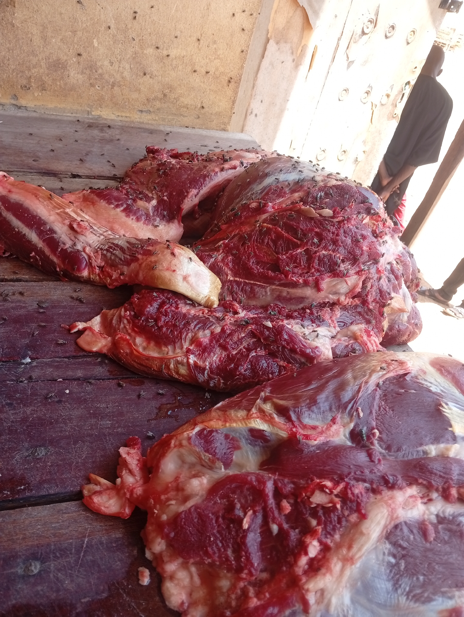 In Barnawa, Nigeria, cuts of raw meat lie on a wooden surface. The meat is heavily covered in flies. A person in dark clothing stands in the background.