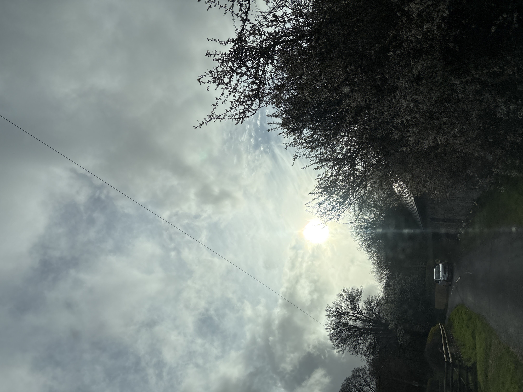 The image shows a partly cloudy sky with the sun shining through. The sun is partially obscured by clouds. In the lower portion of the image is a road flanked by trees and a field. A vehicle is on the road. A power line runs diagonally from the top left. Based on the road configuration, foliage, and vehicle, this scene is possibly located in Shrewsbury, United Kingdom, but without additional landmarks, precise location is difficult to ascertain.