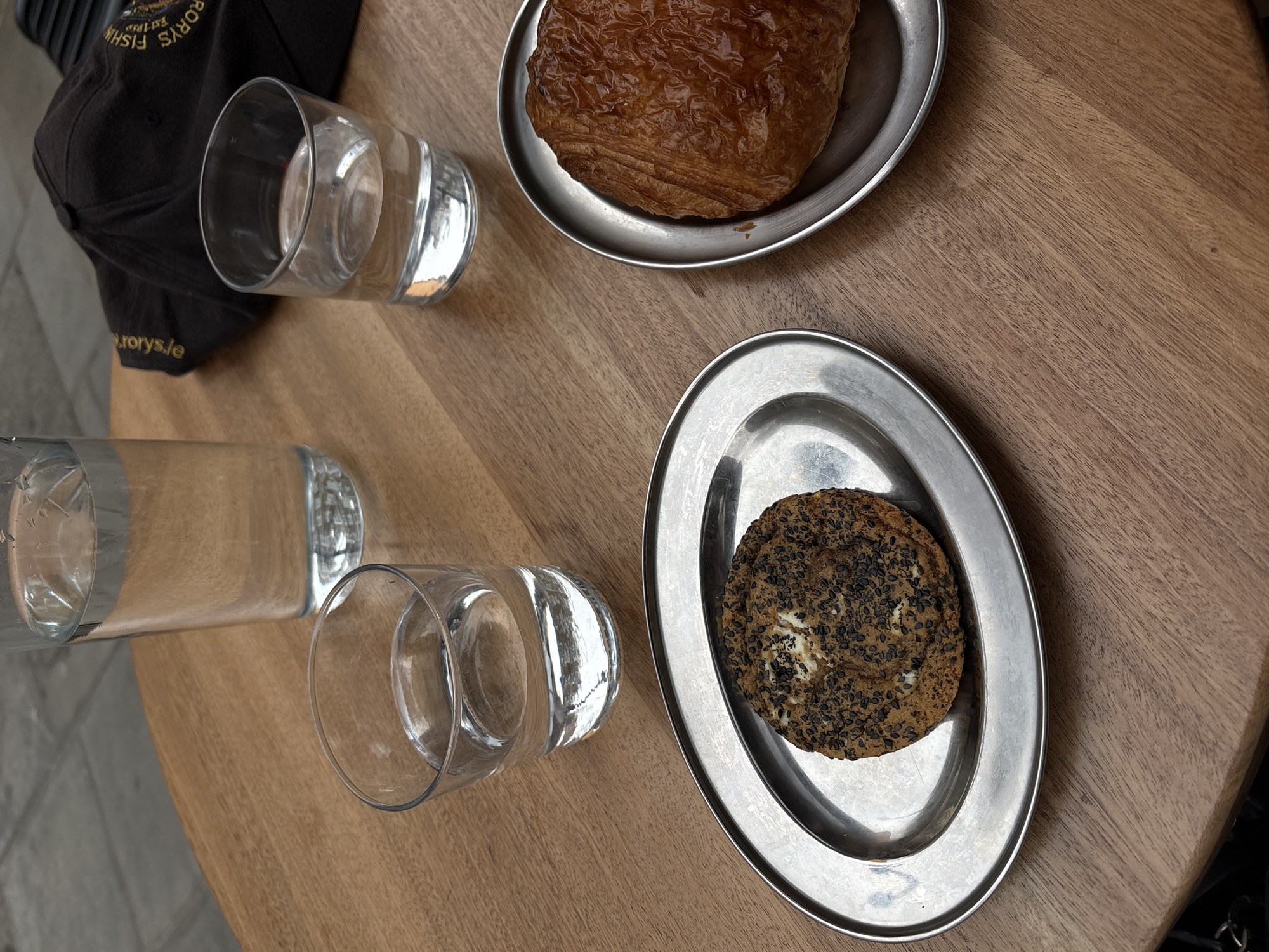 On a light brown wooden table are two small, round metal plates. One plate holds a glazed croissant. The other holds a circular cookie covered in black seeds. Nearby are three glasses, two are upright and appear to contain water, and one is lying on its side. A dark-colored baseball cap is also on the table. The text "rorys.ie" is visible on the cap.