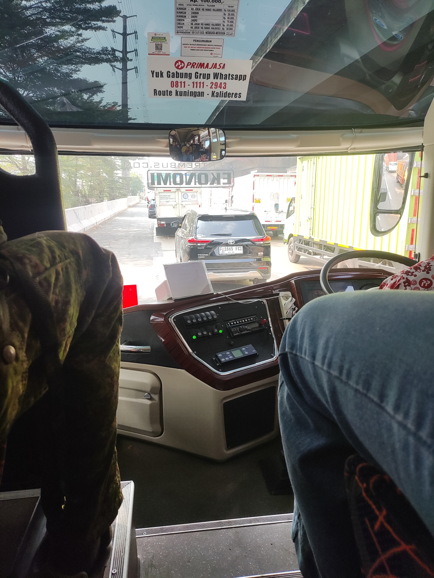 The image is a photograph taken from inside a bus, looking forward through the windshield. The perspective is from the driver's seat. In the foreground, a person wearing a blue denim jacket is visible, their arm and shoulder resting on the steering wheel. The dashboard of the bus is in the center, featuring a complex array of buttons and controls, with wood-grain accents. A dark green fabric, possibly a jacket or seat cover, is visible on the left side of the frame.
Outside the bus, traffic is visible on a road. Several vehicles, including a black SUV with the license plate "F 1865 FCI", a white truck, and a bright yellow truck, are part of a traffic jam. The road appears to be lined with concrete barriers. In the background, trees and overhead power lines can be seen. The sky is overcast.
On the windshield, there are several stickers. One prominent sticker displays the text "PRIMAJASA" and "Yuk Gabung Grup WhatsApp 0811-1111-2943 Route Kuningan - Kalideres". Another sticker has a QR code and the text "PRIMAJASA". A smaller sticker above it contains a list of routes and prices. A mirror is positioned above the dashboard, reflecting a group of passengers inside the bus.