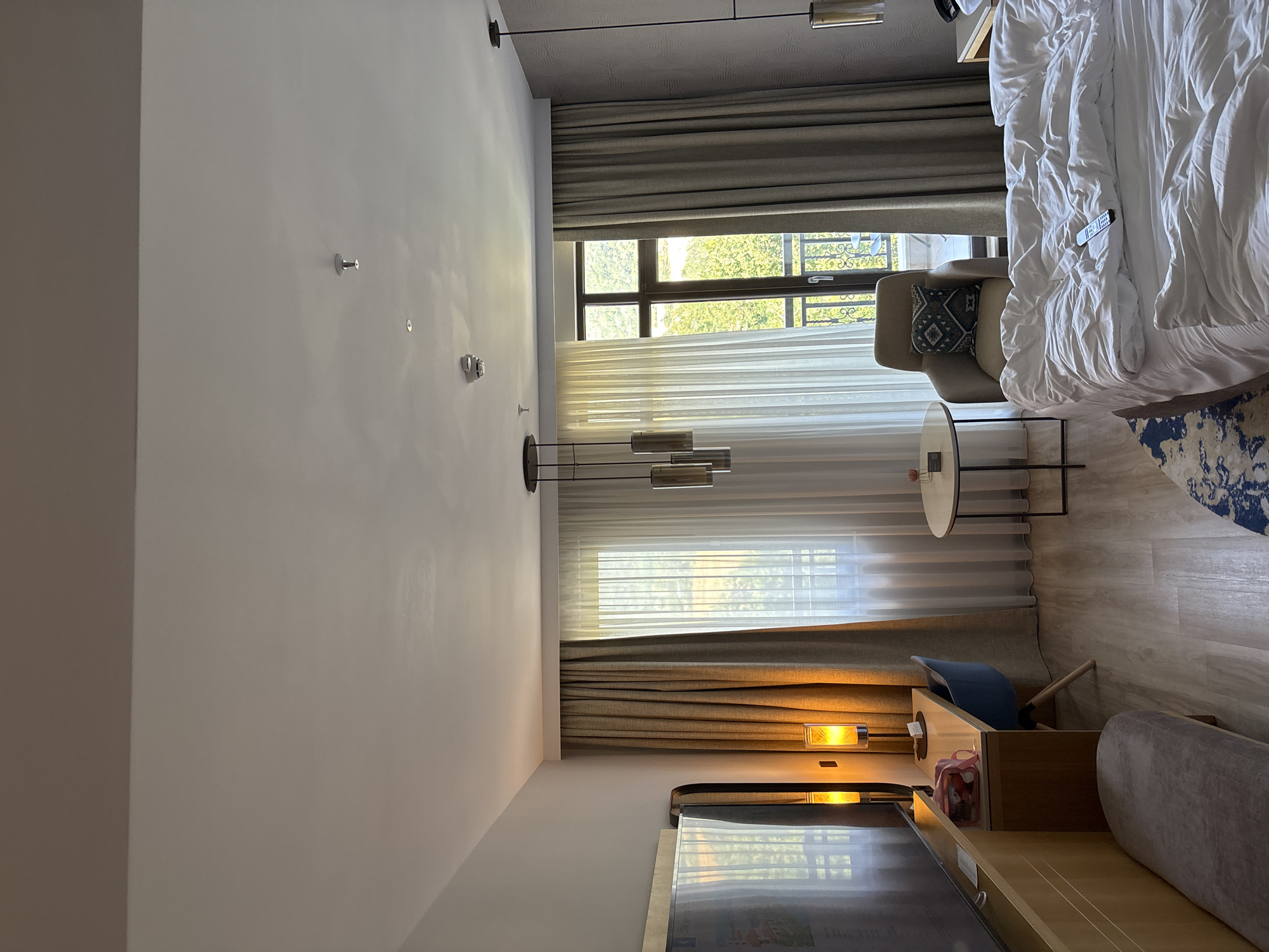 The image depicts a hotel room, likely located in Razliv, Bulgaria, with a focus on the interior furnishings and arrangement. In the foreground, a rumpled bed with white linens dominates the right side of the frame, with a remote control resting on the duvet. To the left, a desk area is visible, featuring a light-colored desk with a blue chair, a desk lamp casting a warm glow, and a small pink bag. Adjacent to the desk is a large flat-screen television mounted on a wooden fixture, with a padded bench situated in front of it. The room has large windows covered by both sheer white curtains and heavier beige drapes, which reveal a glimpse of greenery and a balcony beyond. A small round side table with a minimalist design stands near an armchair, which is adorned with a patterned cushion. The ceiling shows light fixtures and what appear to be smoke detectors. The overall impression is that of a well-appointed hotel room, possibly in a resort setting given the view of trees.