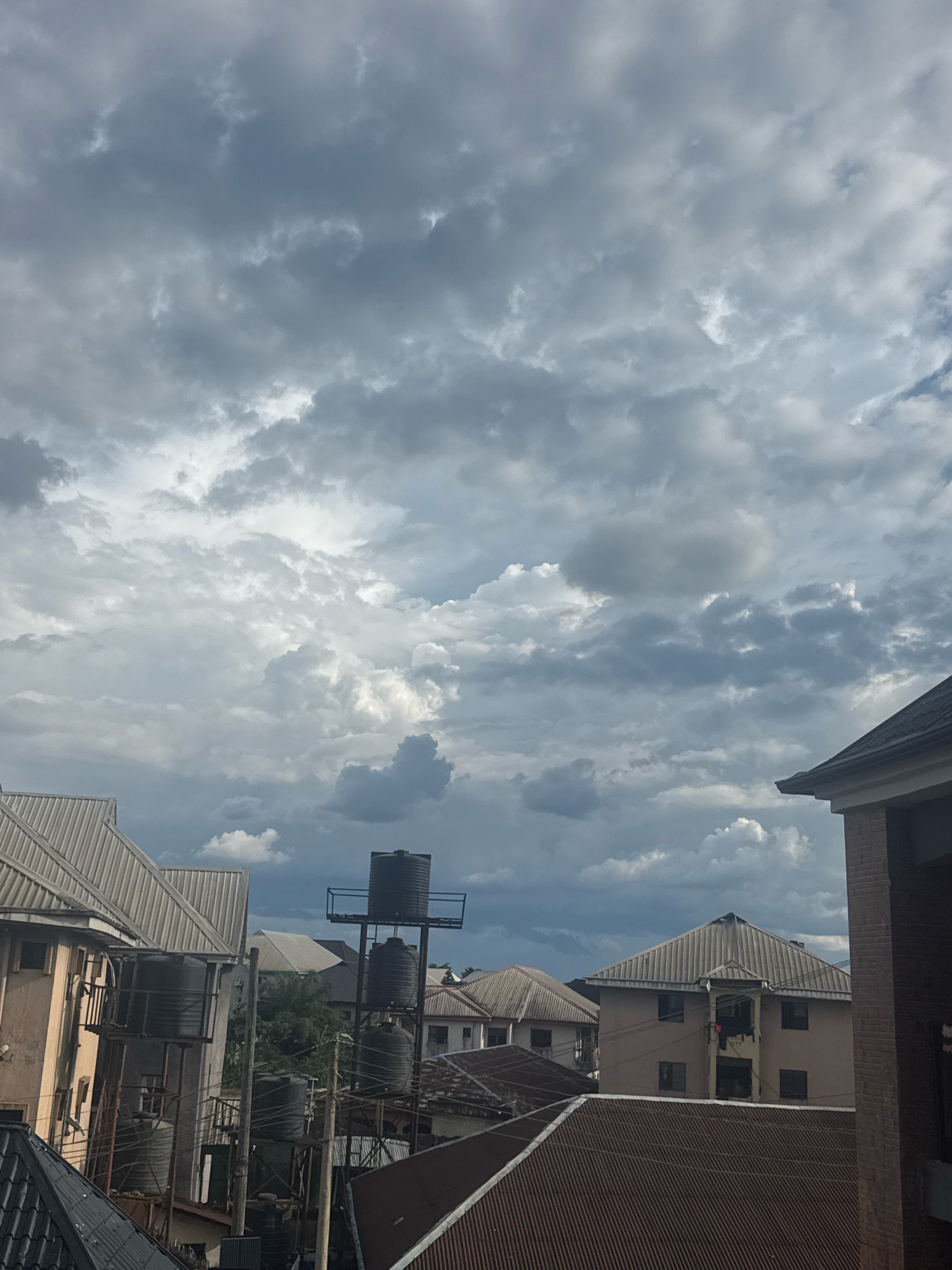 The image shows a view of rooftops and buildings in Ihiagwa, Nigeria, under a cloudy sky. Several residential buildings with corrugated metal roofs are visible, some with water tanks mounted on stands. One building in the foreground on the left has a light-colored facade with multiple windows and balconies. To the right, another building with a brown brick facade and a dark, sloped roof is partially visible. In the background, more buildings with similarly styled roofs can be seen, interspersed with trees. A tall structure supporting three large black water tanks is a prominent feature in the mid-ground. The sky is filled with a mixture of dark and light gray clouds, suggesting overcast or potentially stormy weather. There are no people or animals visible in the frame. The scene appears to be an urban or semi-urban residential area.