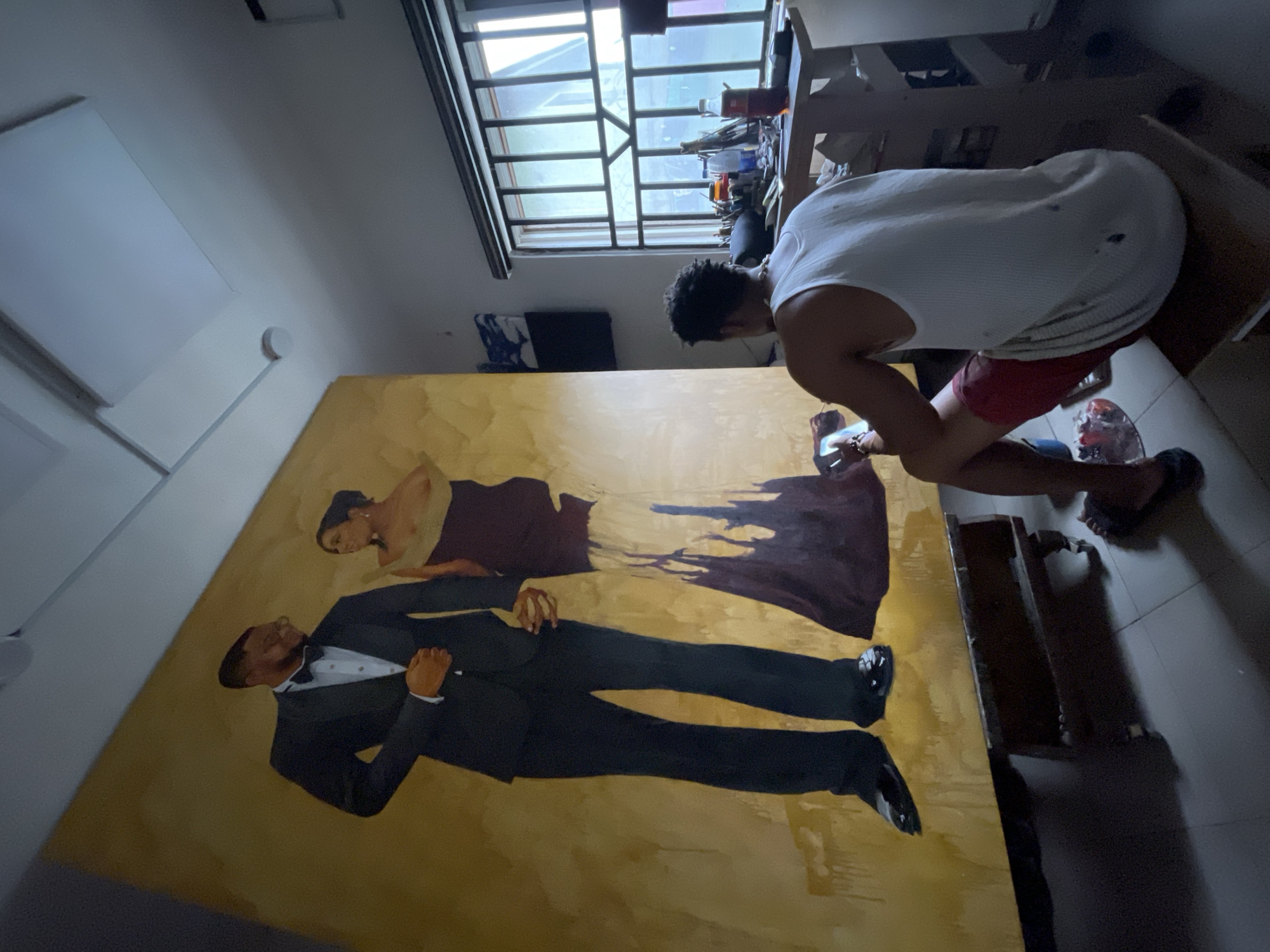 In a room that appears to be a studio, located in the city of Ado, Nigeria, a large, unfinished oil painting of a couple stands on an easel. The painting, set against a golden background, depicts a man in a tuxedo and a woman in an elegant, flowing gown. A young man, seated on a stool, is actively working on the painting. He is wearing a white tank top and red shorts. His right hand holds a palette with paint, and his left hand is holding a brush, with which he is applying paint to the canvas. The studio is lit by natural light filtering through a window covered with security bars. Various art supplies, including brushes, bottles, and a palette, are visible on a nearby table. The overall atmosphere suggests a dedicated artist engrossed in their work within a functional, albeit somewhat cluttered, studio space.